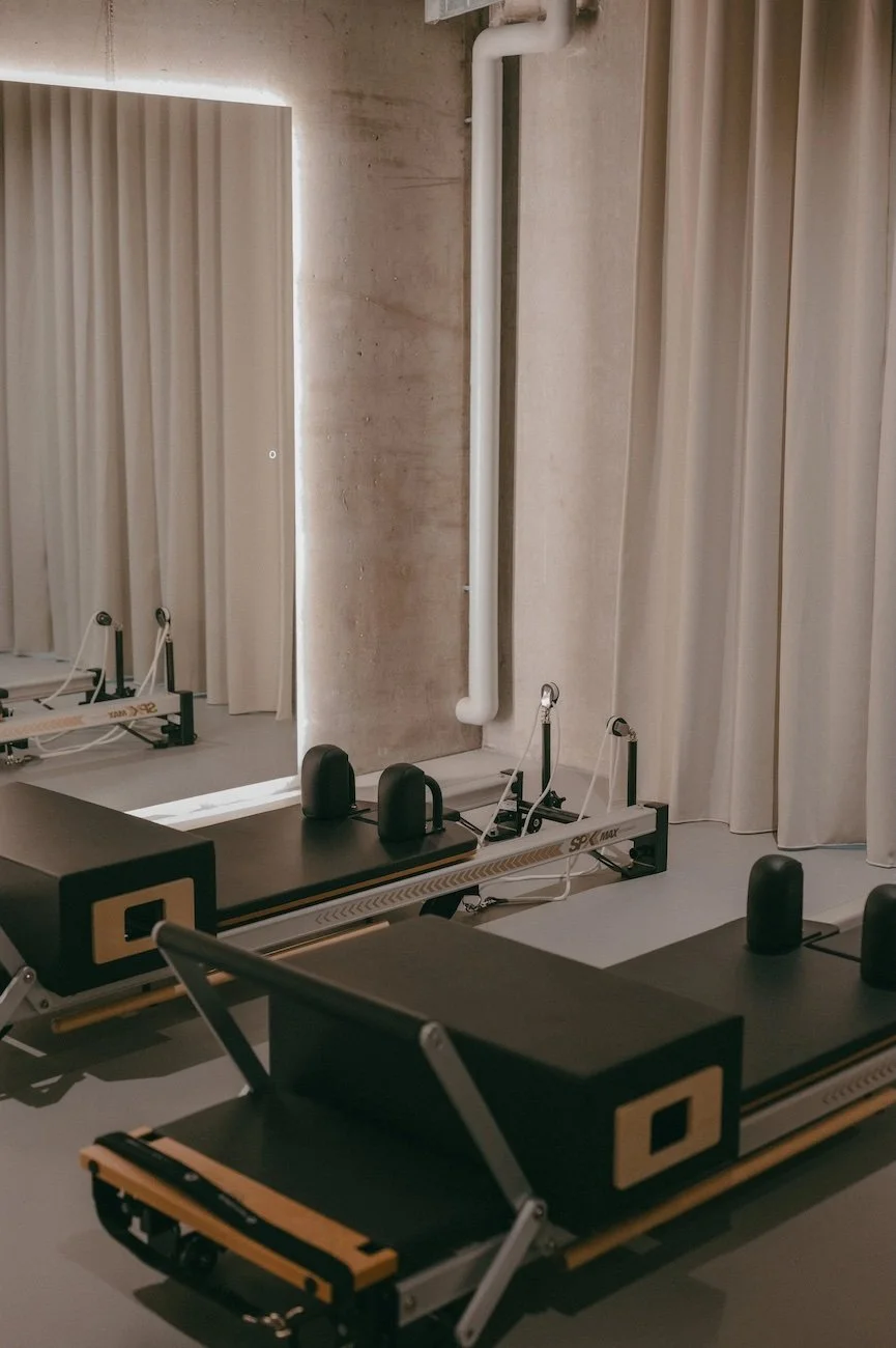 Two Pilates reformer machines in a studio with beige curtains and a concrete wall.