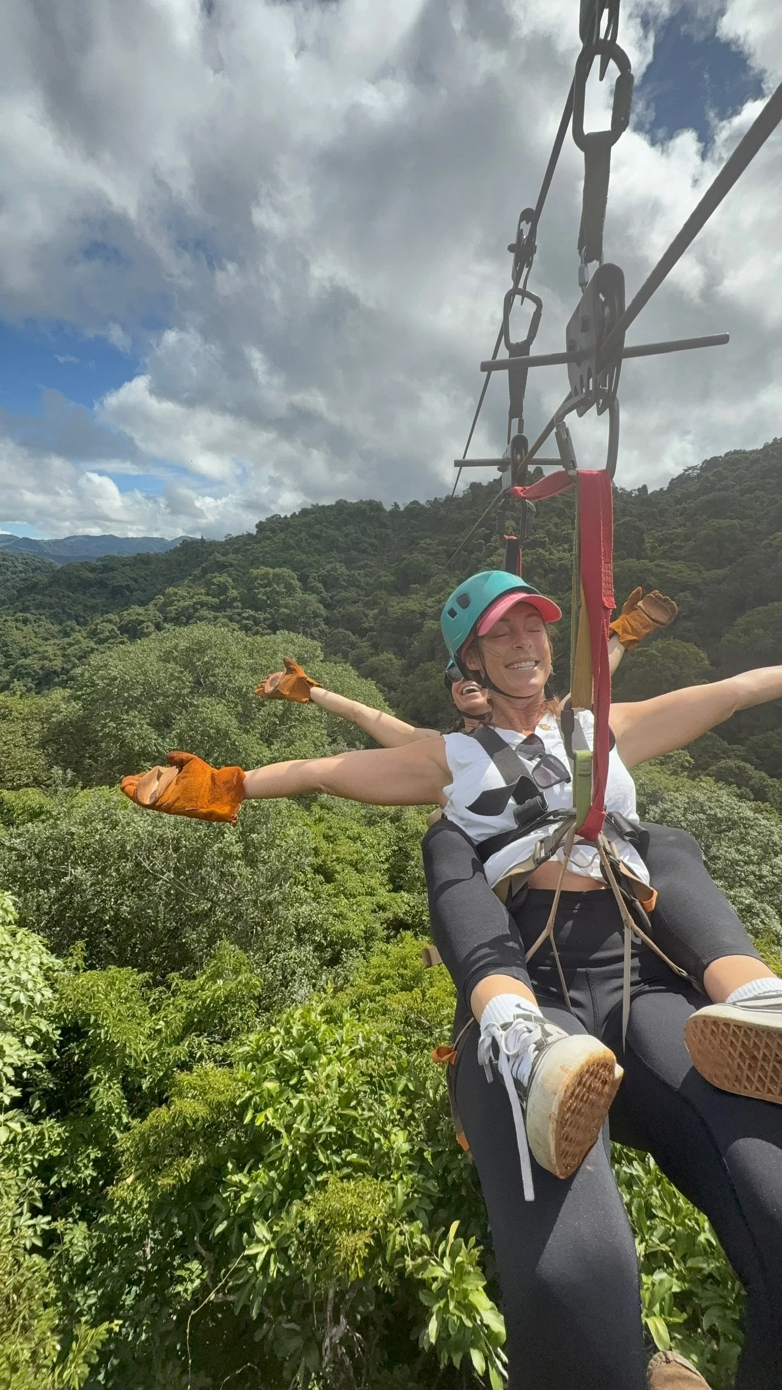 A woman smiling on a zip line with her arms outstretched, in a lush green forested area.