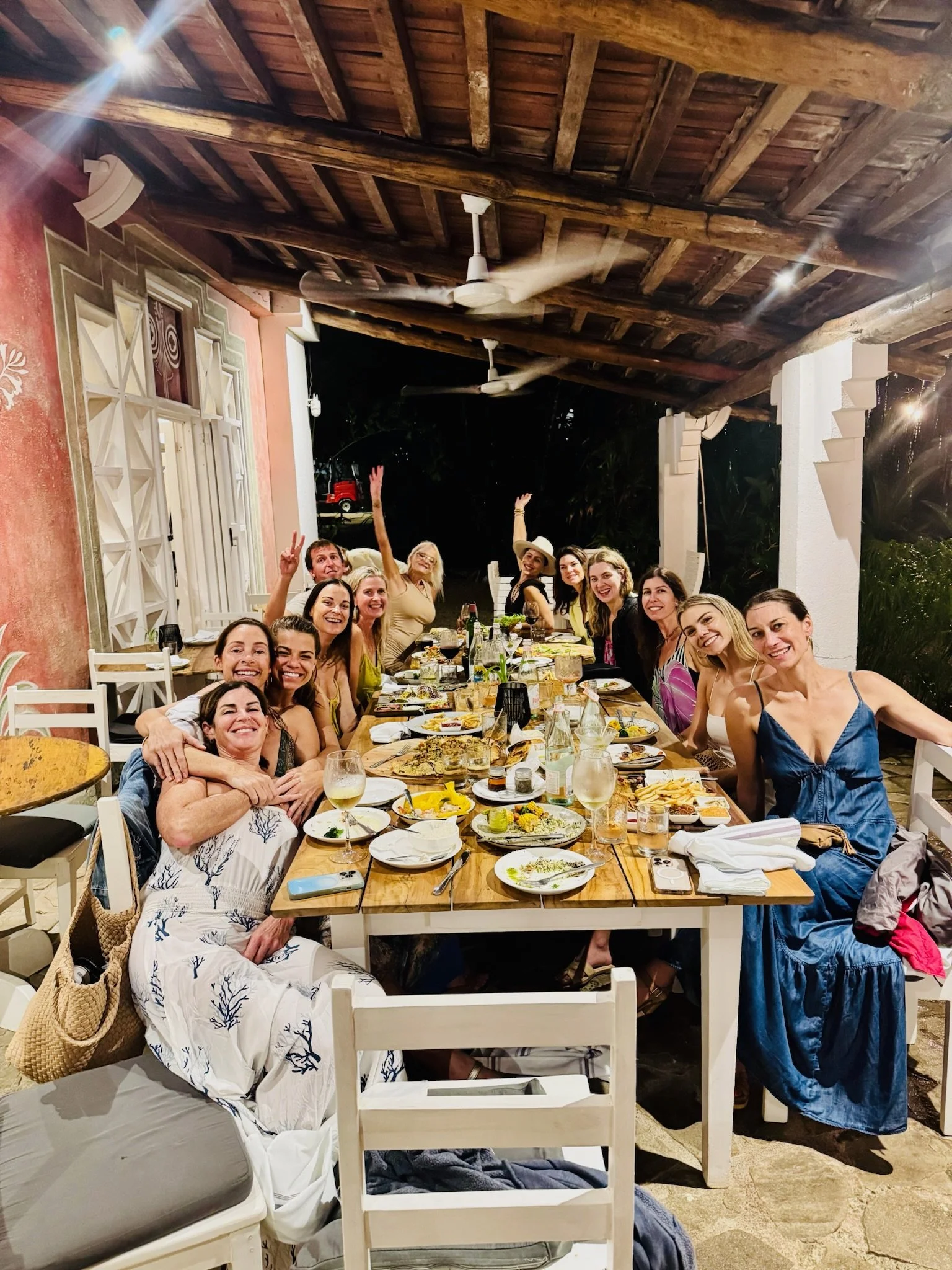 A group of women gather around a long dinner table outdoors at night, enjoying food and drinks. The setting features a wooden ceiling with fans, and the women are smiling and celebrating.