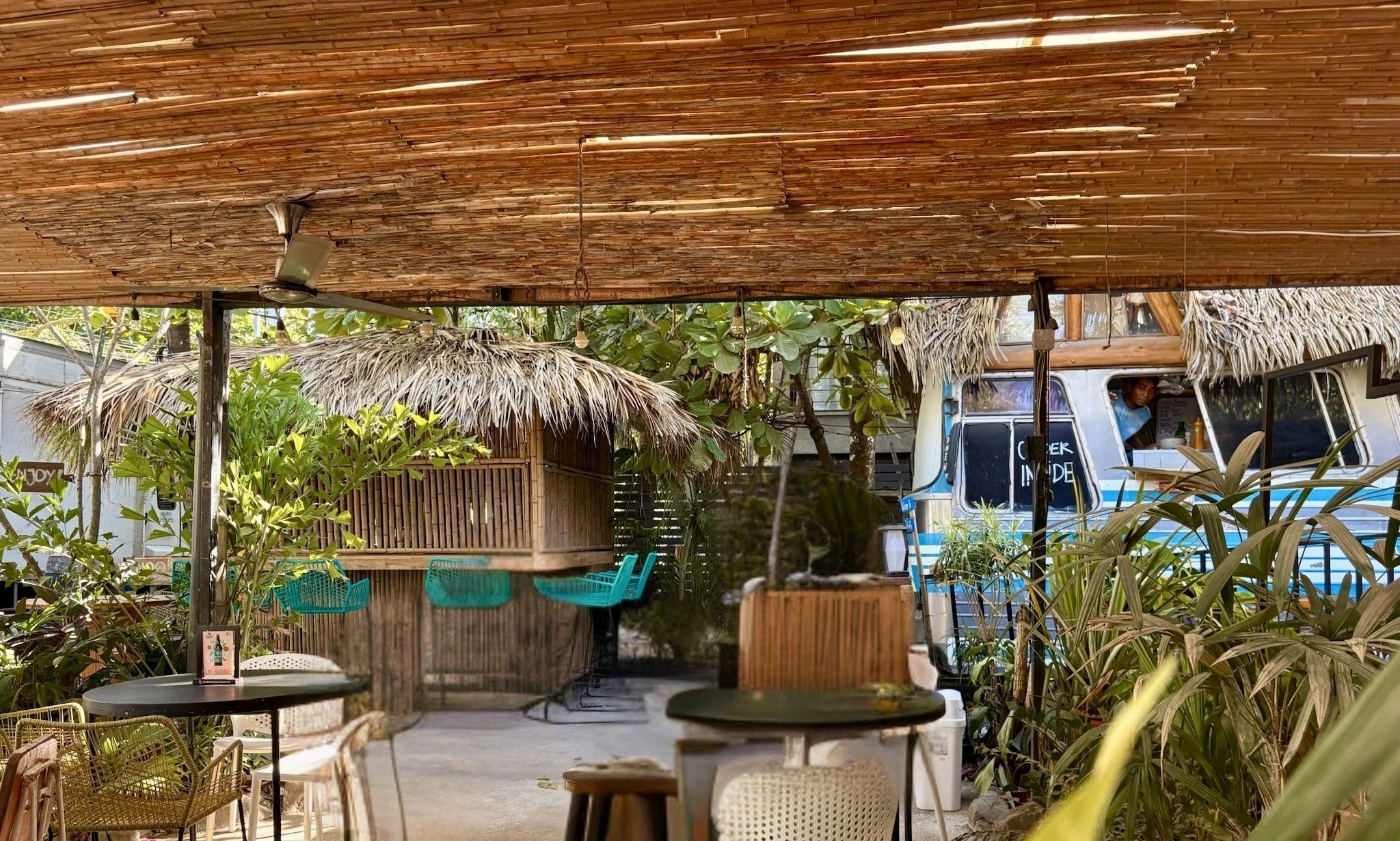 Outdoor cafe with bamboo and thatch roof, surrounded by lush greenery, with a vintage food truck in the background and seating areas with chairs and tables.