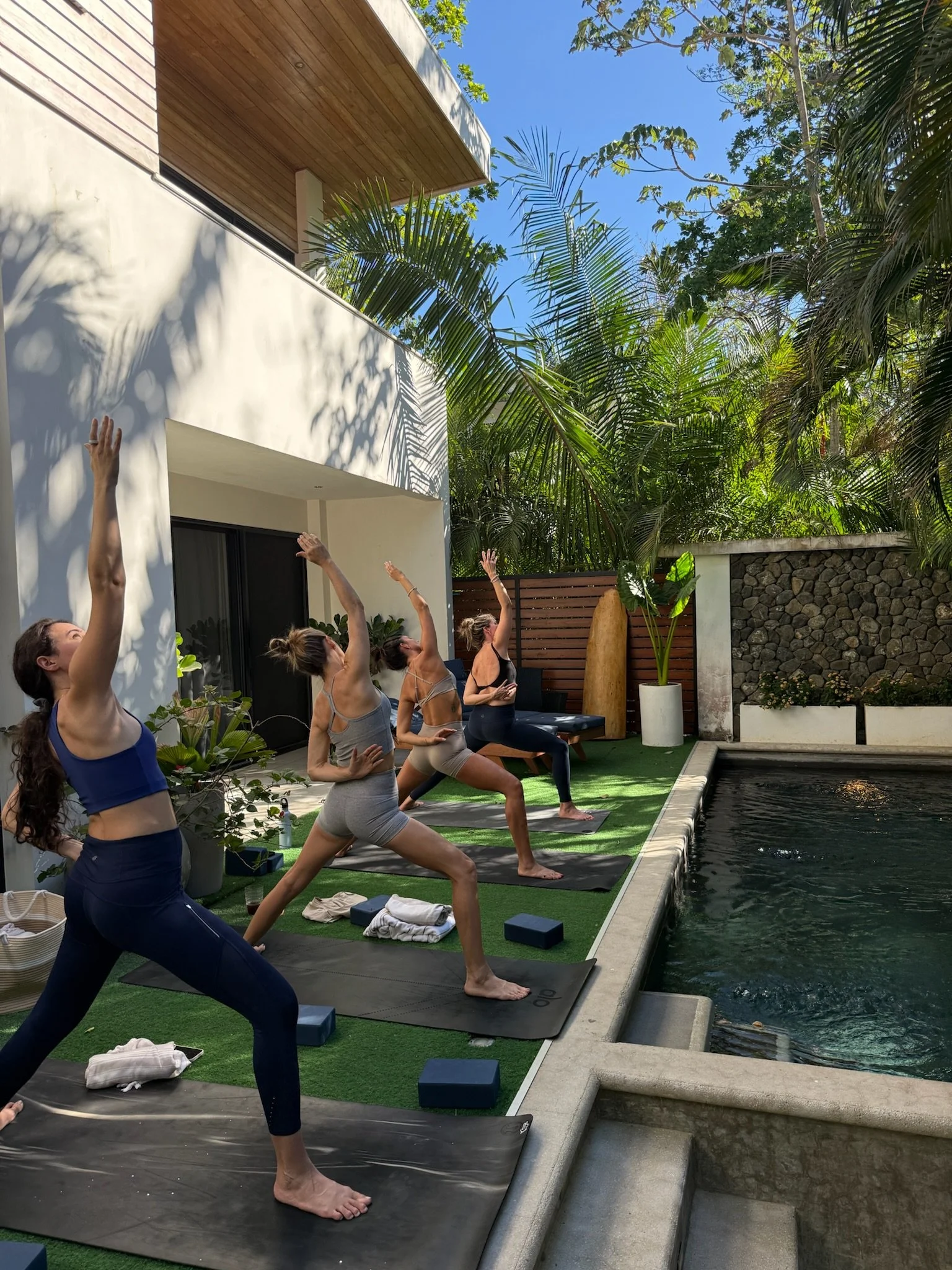 Group of women practicing yoga outdoors near a pool, sitting on mats in seated meditation poses with closed eyes, surrounded by lush greenery.