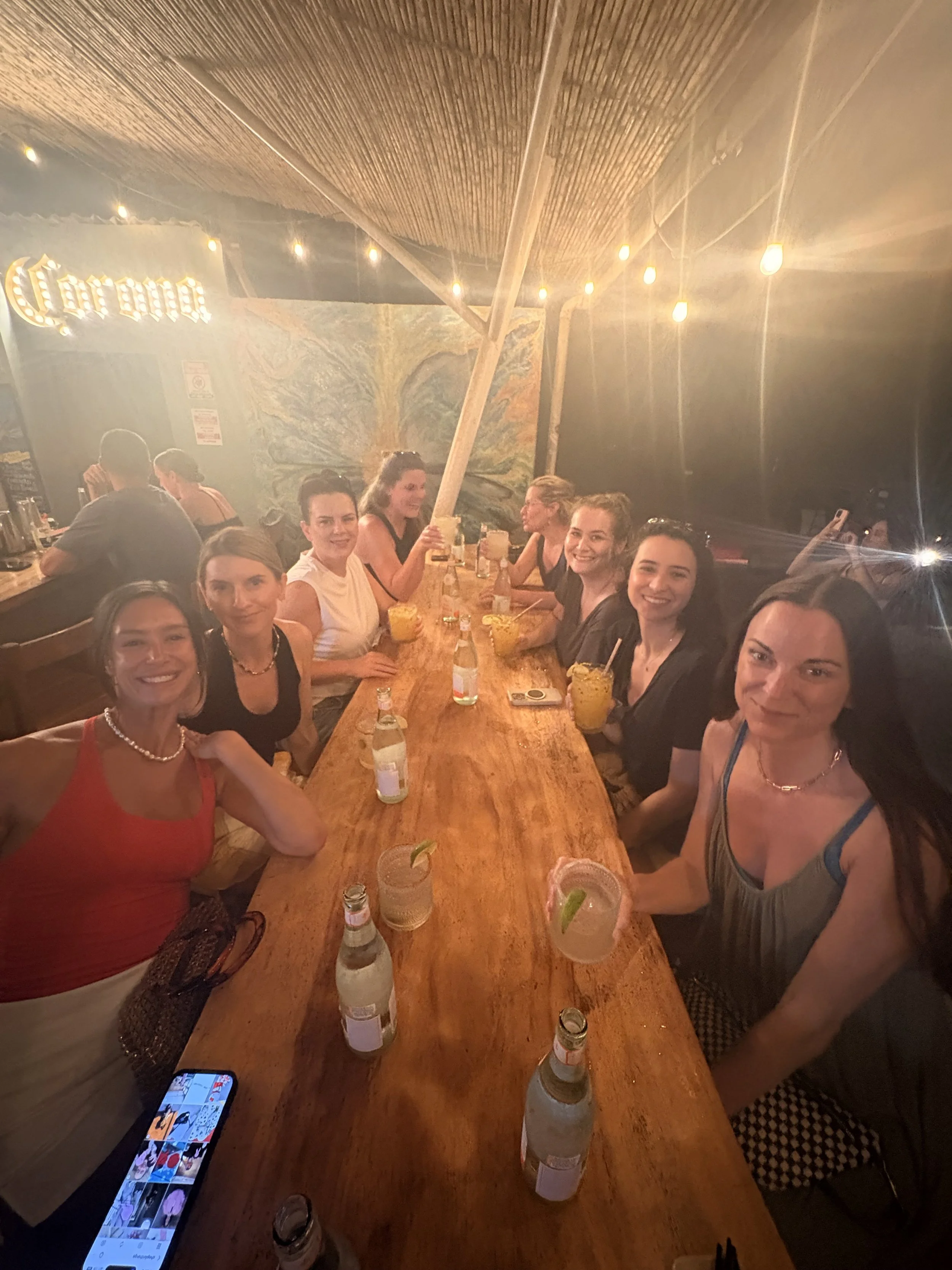 A group of women gather around a long dinner table outdoors at night, enjoying food and drinks. The setting features a wooden ceiling with fans, and the women are smiling and celebrating.