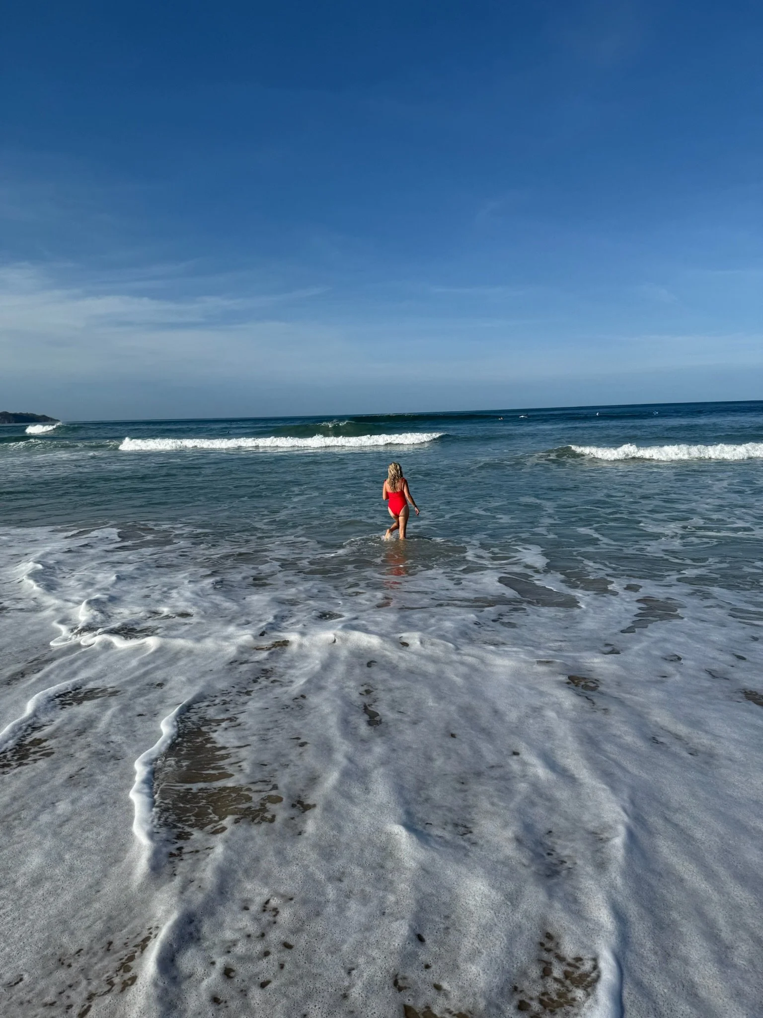 A woman in a red swimsuit wading into the ocean waves on a sunny day, with a clear blue sky overhead.