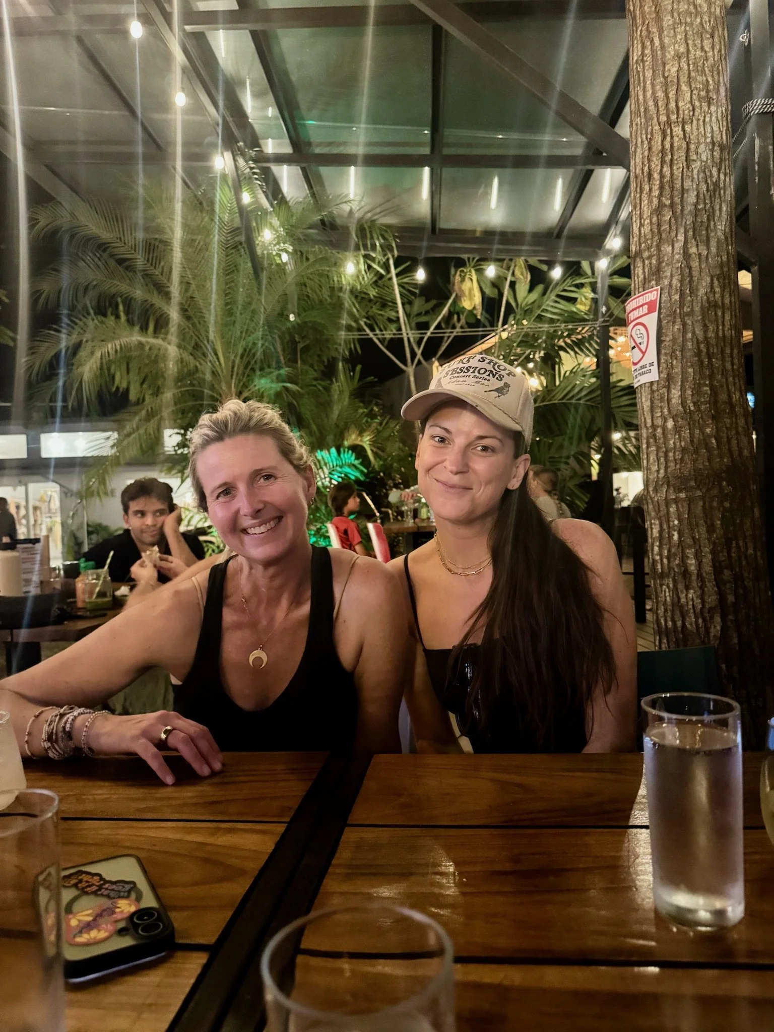 Two smiling women sitting at a wooden table in an outdoor restaurant at night, with one wearing a black top and cap, and the other wearing a black top with jewelry, surrounded by lush green plants and string lights.