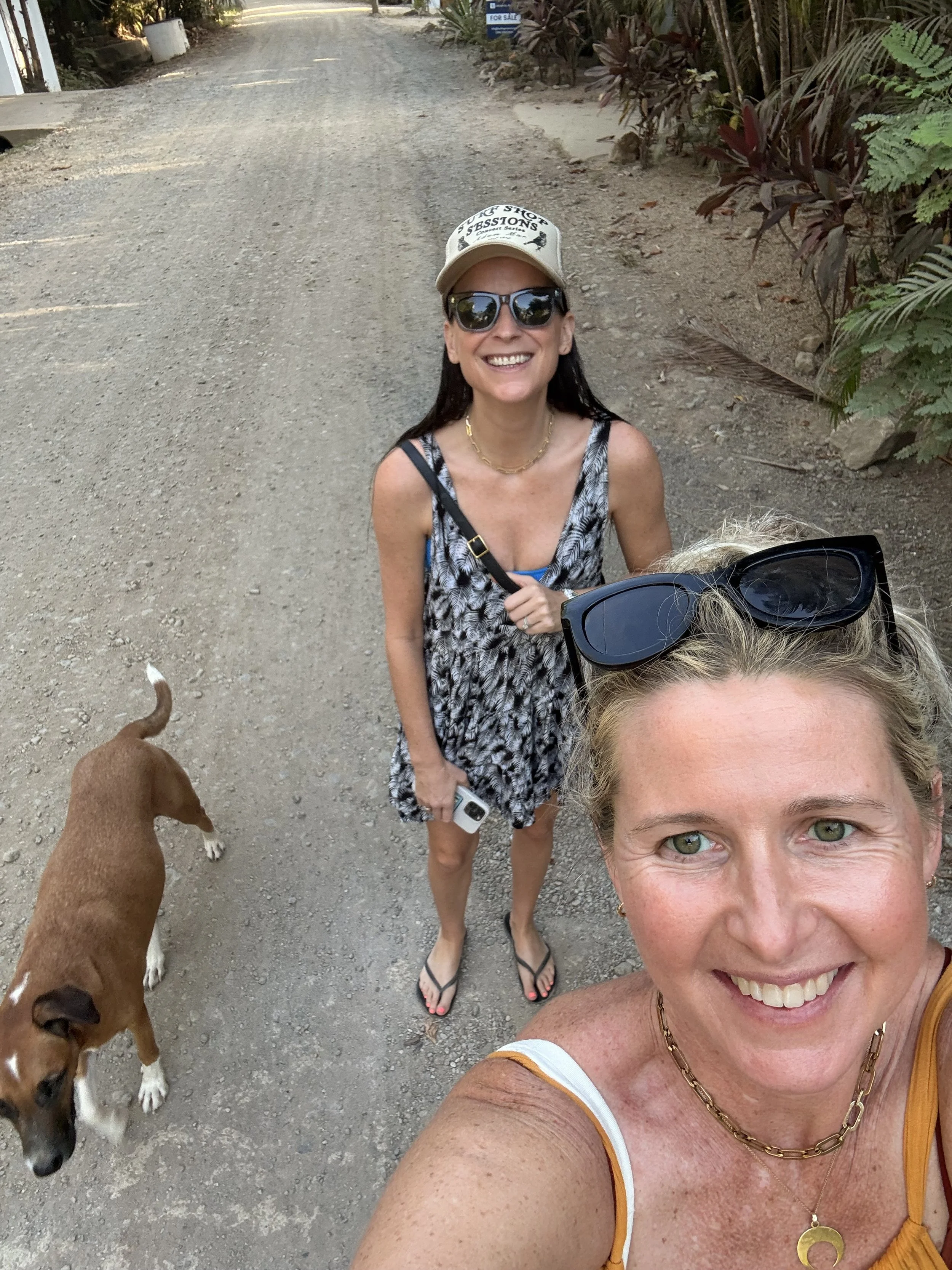 Two women taking a selfie on a dirt road with a dog, surrounded by plants and trees.