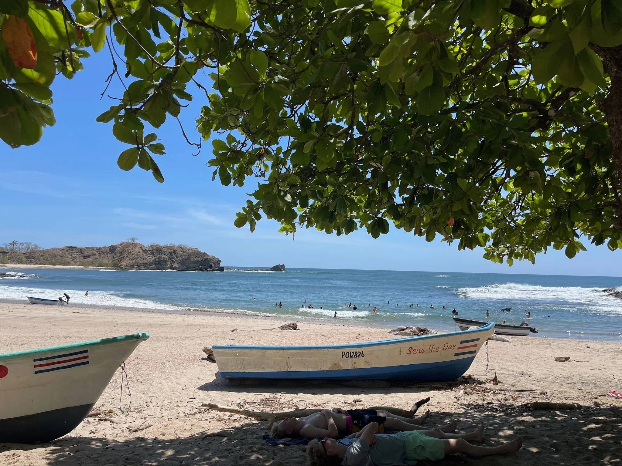 Scenic beach with boats on sandy shore, people swimming and relaxing in the water, and lush green trees overhead, with rocky formations in the distance.