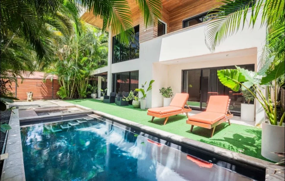 Modern backyard with swimming pool, two pink lounge chairs, potted plants, tropical trees, and a contemporary house with large windows and a covered patio.