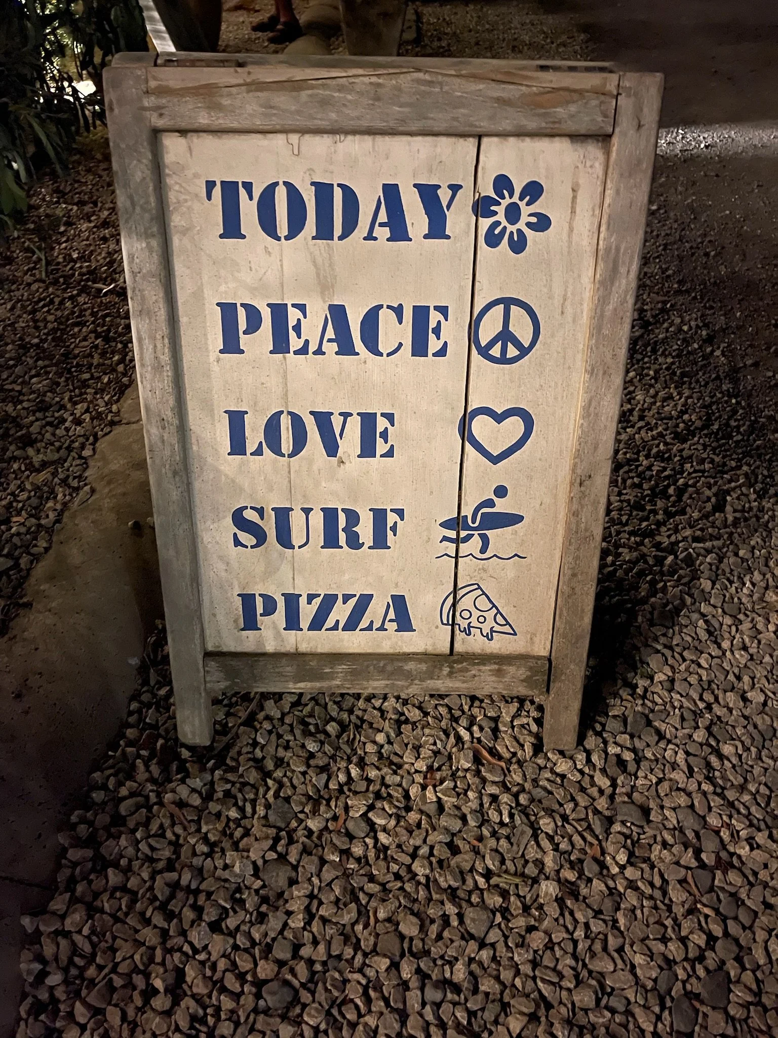 A wooden signboard with the words 'Today Peace Love Surf Pizza' in blue text, accompanied by symbols of a flower, peace sign, heart, surfboard, and pizza slice.