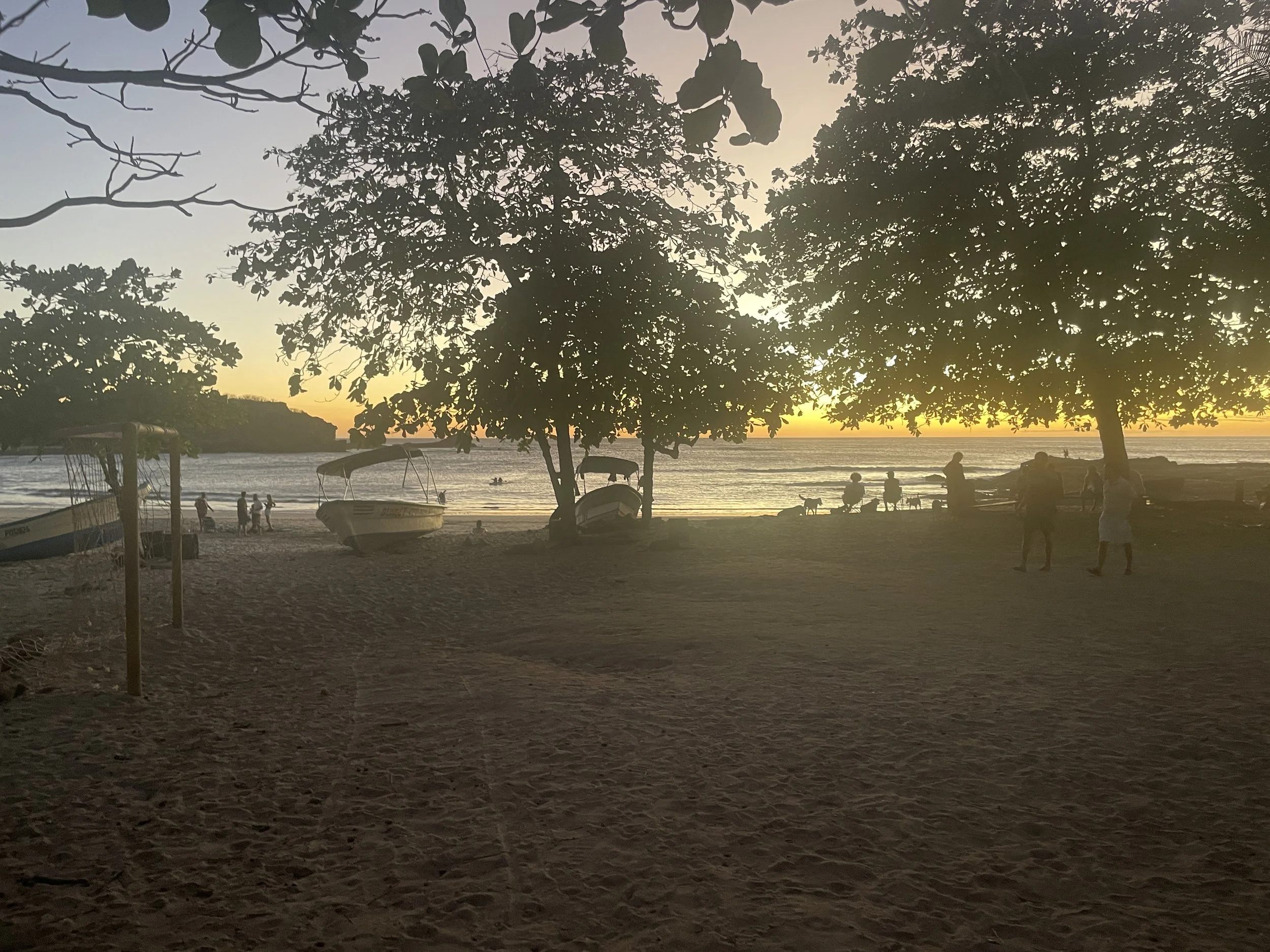 A beach at sunset with boats on the sand and trees silhouetted against the sky. People are walking, sitting, and enjoying the scene near the water.