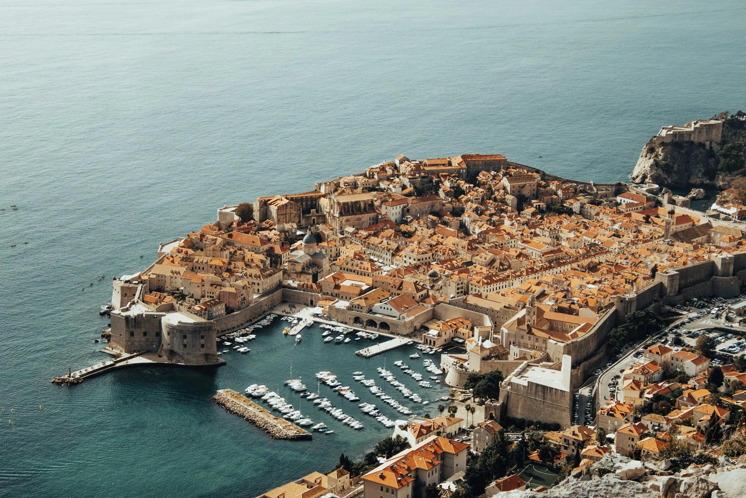 Aerial view of old city with red-tiled roofs, surrounded by stone walls, near a harbor with boats, on a sunny day in a coastal area.