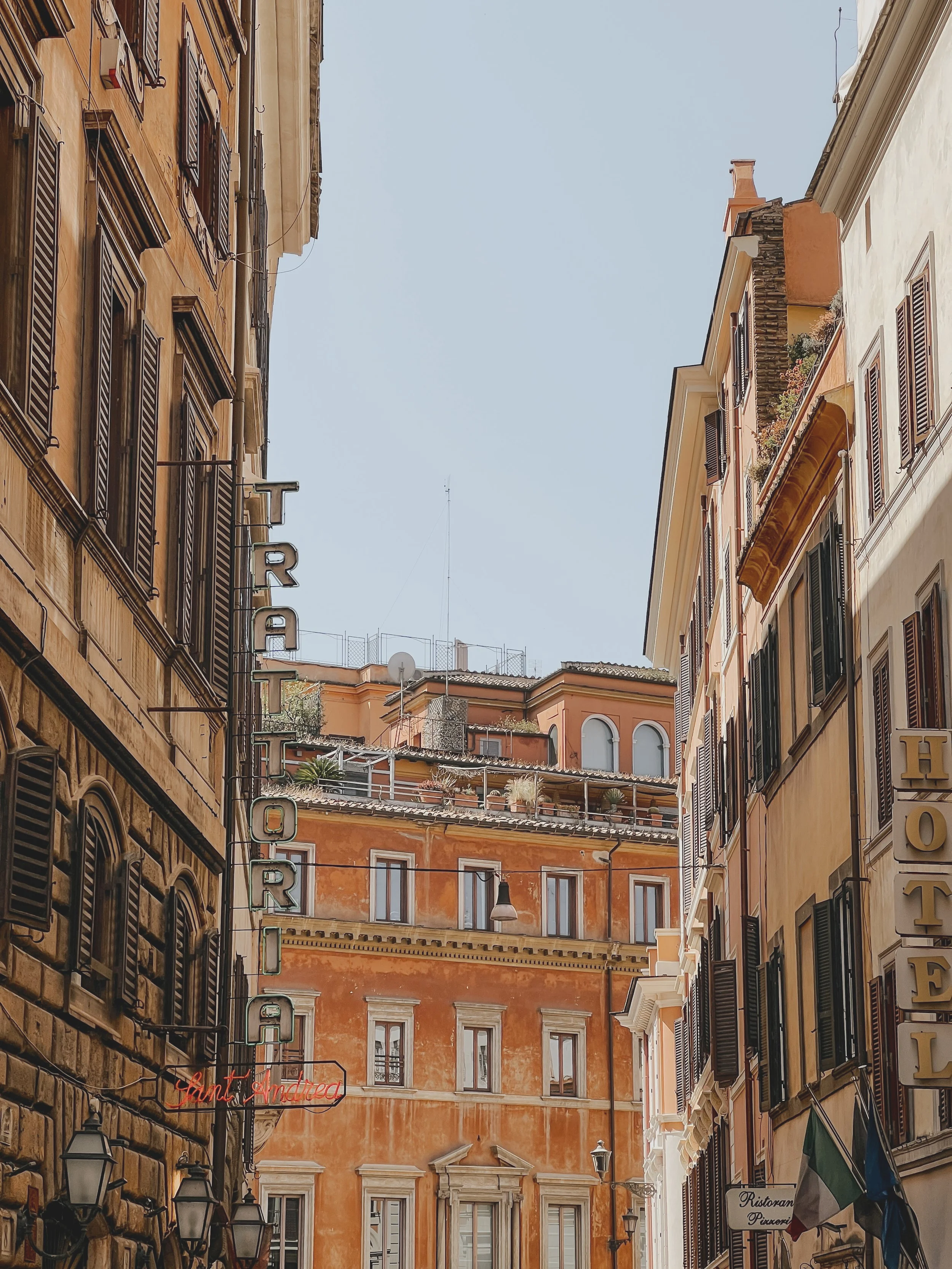 Narrow street in Italy with colorful old buildings, signs for a hotel, restaurant, and a pizzeria, and a clear blue sky overhead.