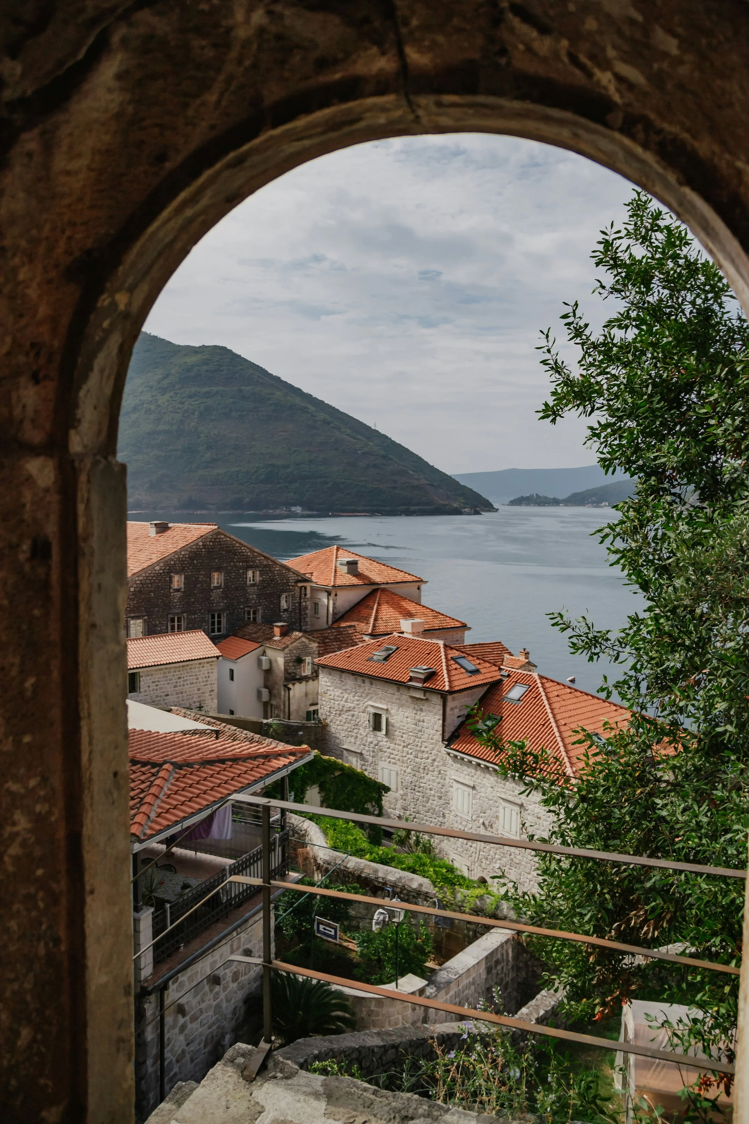 View through a stone archway showing houses with red tile roofs, a body of water, a mountain in the background, and partly cloudy sky.