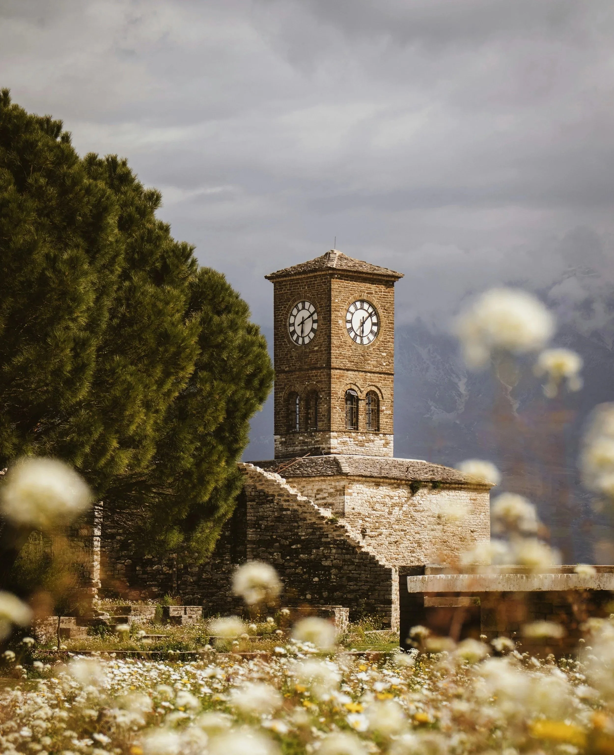 An old stone clock tower with two clock faces, surrounded by greenery and wildflowers, under a cloudy sky.