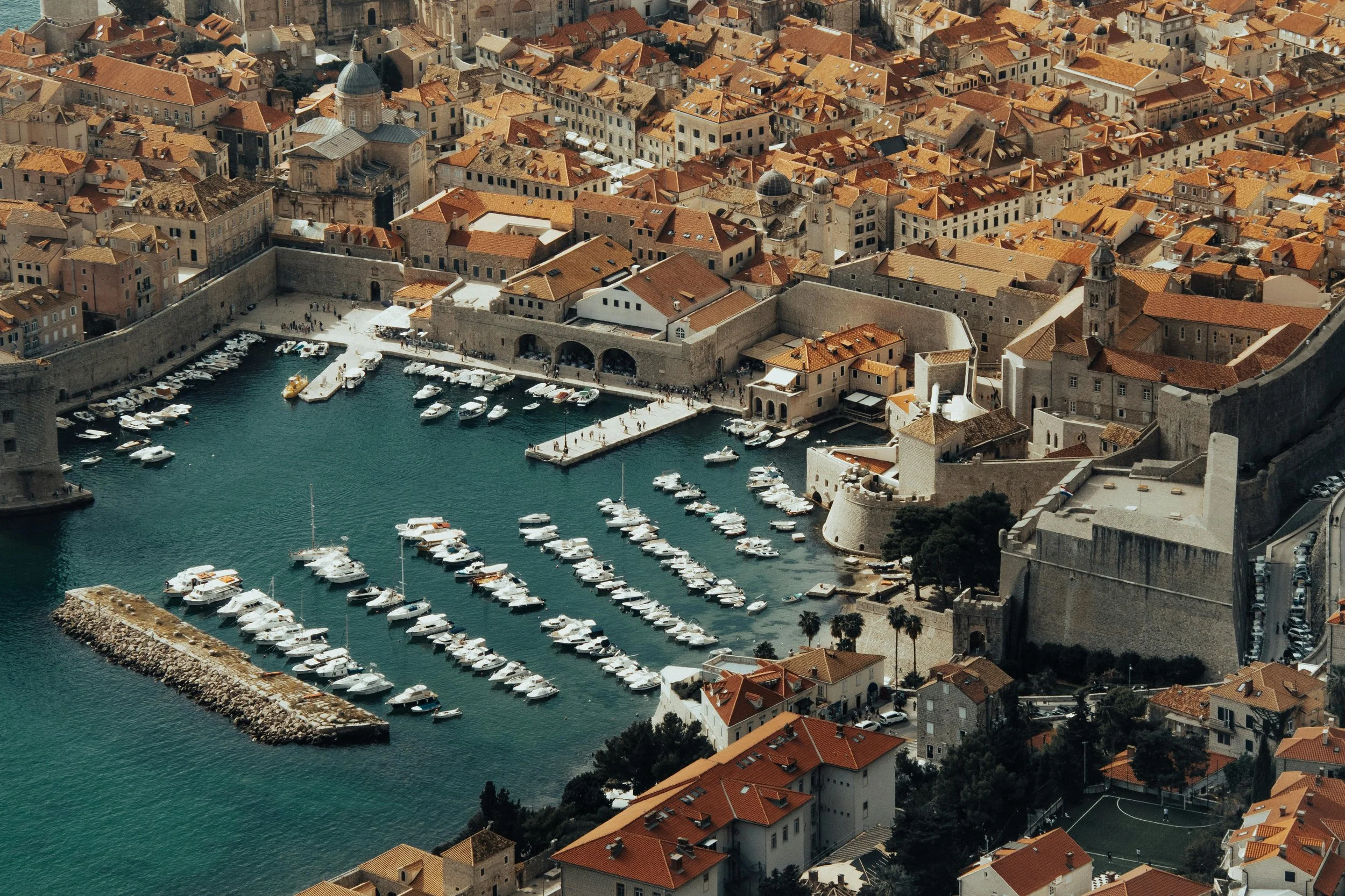 An aerial view of a historic coastal city with a fortified stone wall, a harbor filled with docked boats, and terracotta-roofed buildings.
