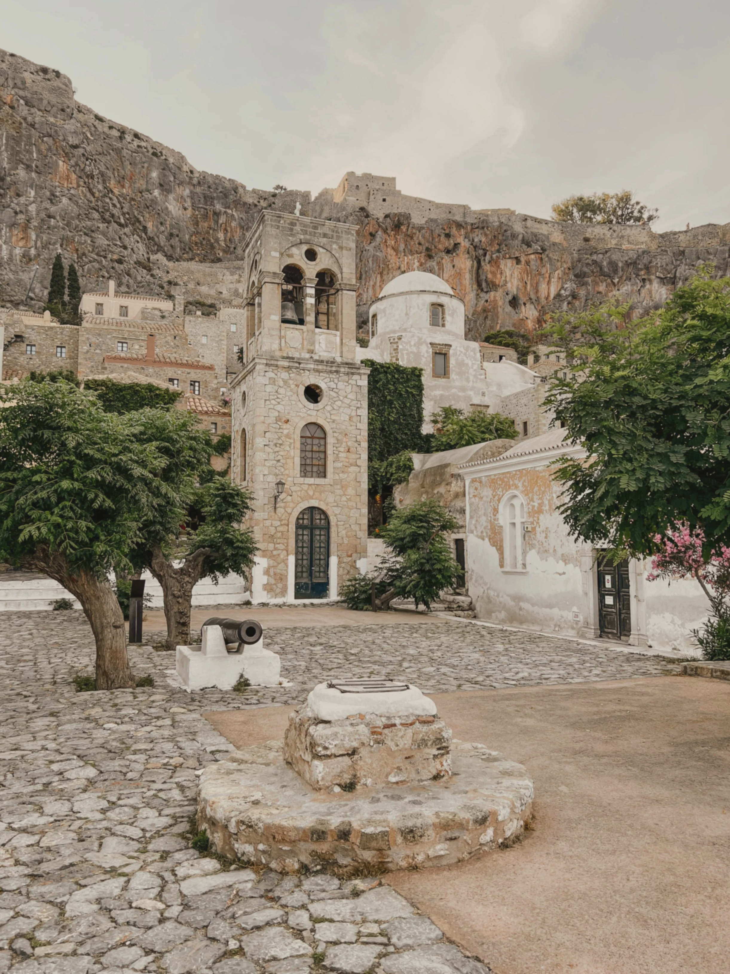 Historic stone church with a bell tower, set against a rocky hillside with old buildings. Trees and a cobblestone courtyard in the foreground.