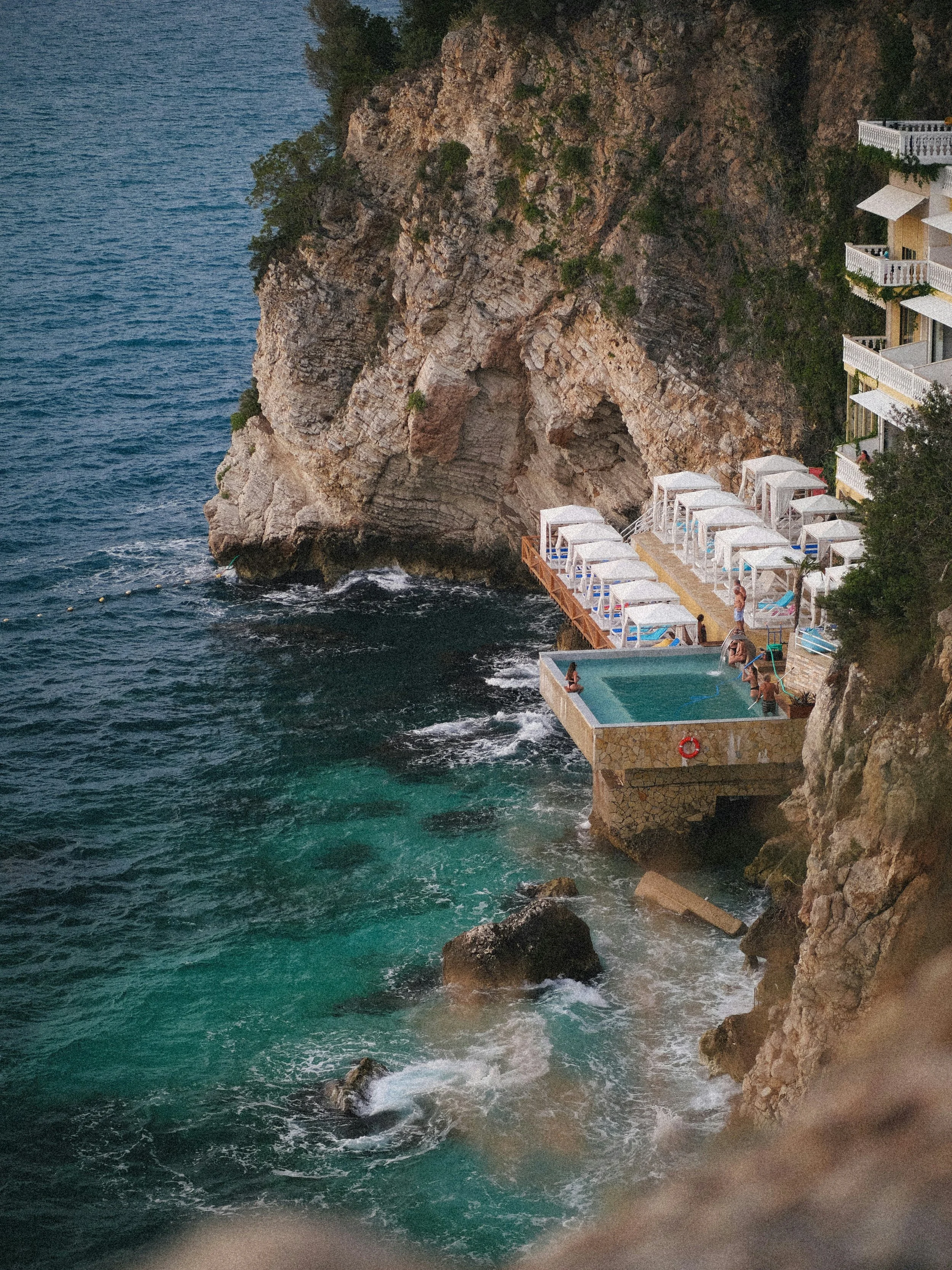 A cliffside resort with a swimming pool and white cabanas overlooking the ocean, with people relaxing and enjoying the view.