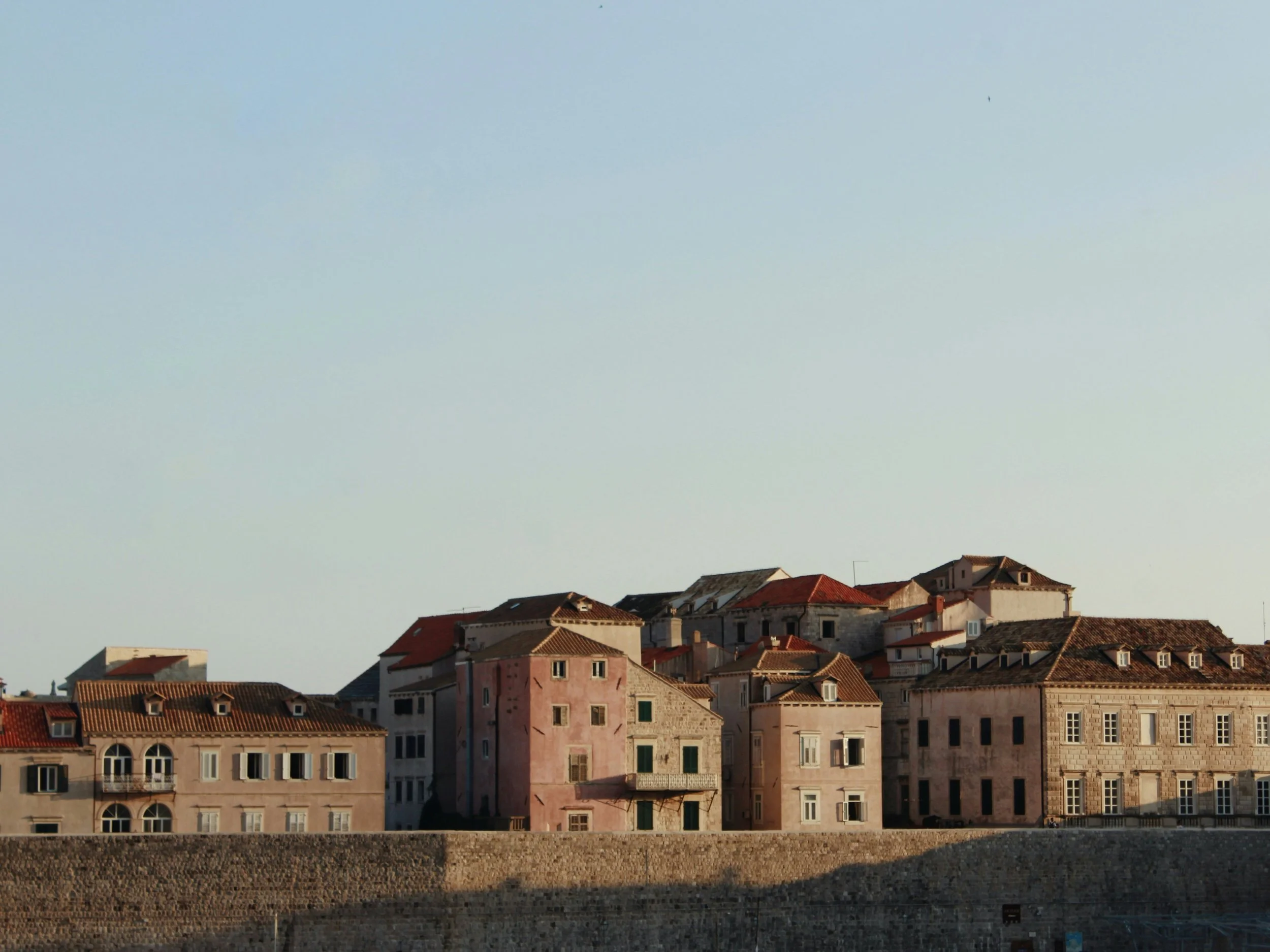 A row of old European-style buildings with various window styles and tiled roofs, situated along a stone wall under a clear sky.