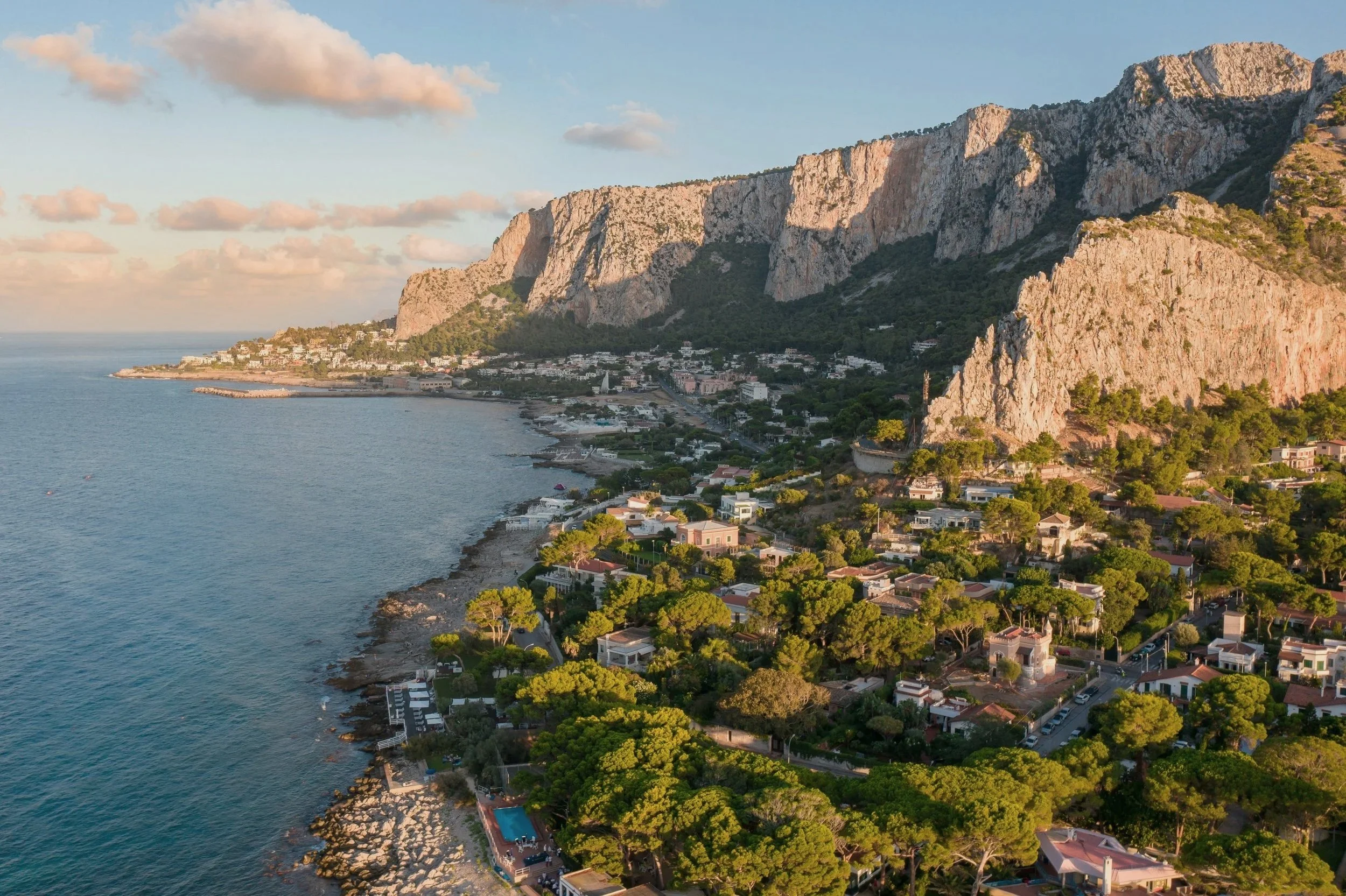 Aerial view of a coastal town with houses and lush green trees, backed by steep rocky cliffs, during sunset.