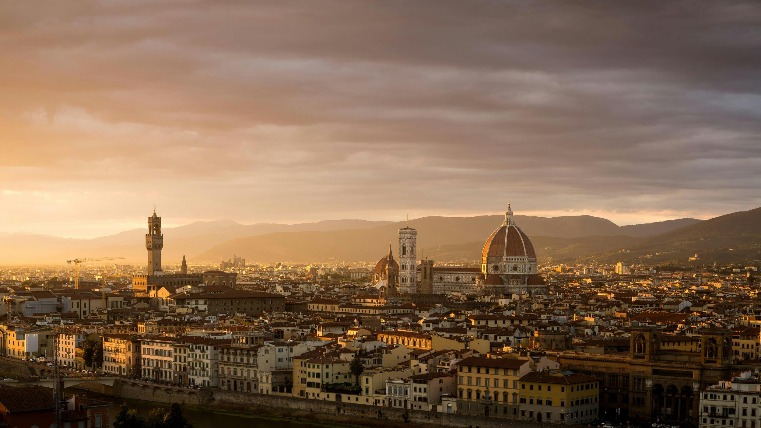 A cityscape of Florence, Italy at sunset showcasing the Florence Cathedral with its iconic dome, surrounded by historic buildings, mountains in the background, and a partly cloudy sky.
