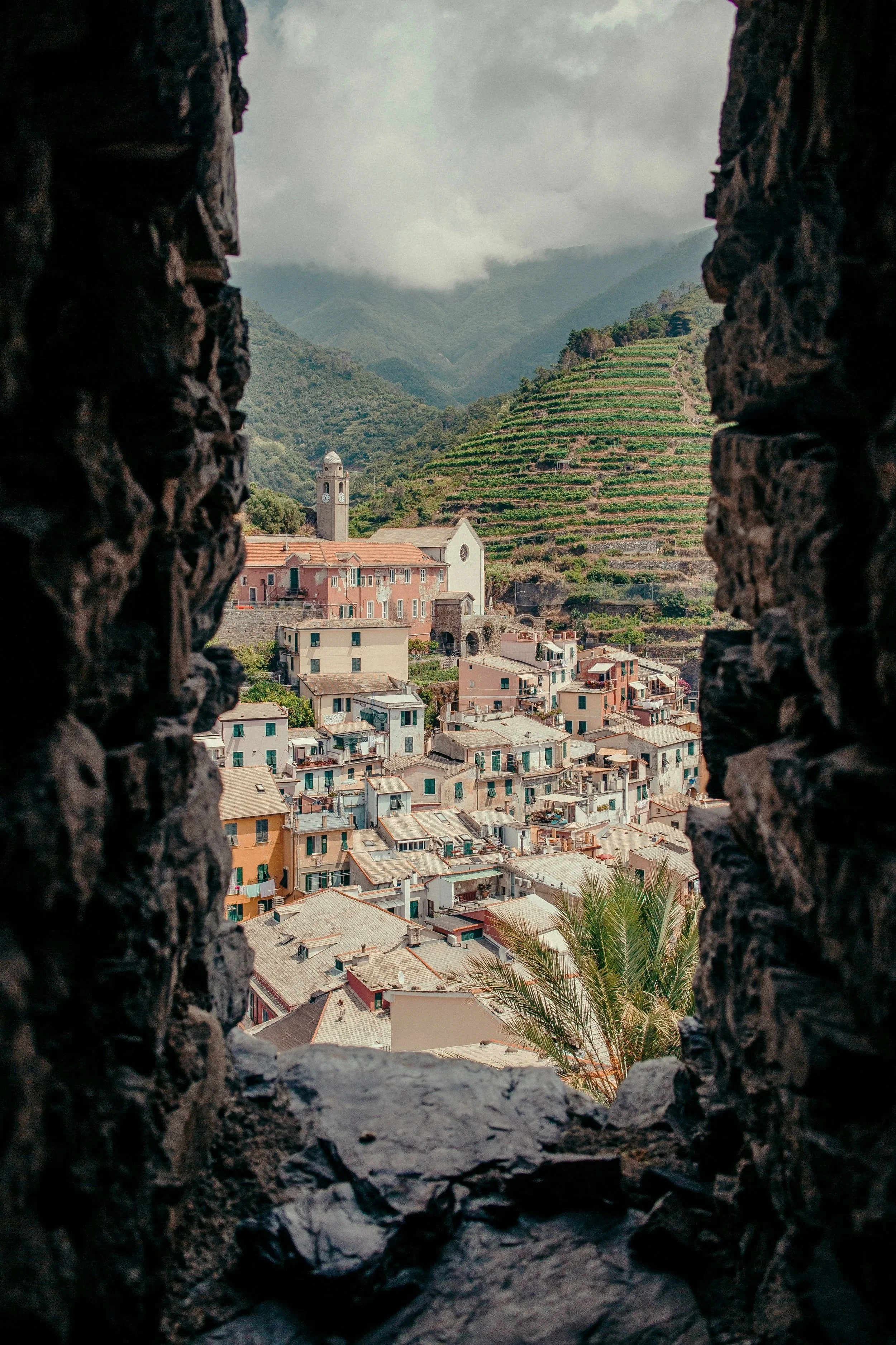 View of a hillside town with colorful buildings, a church with a clock tower, terraced green hills, and mountains in the background, seen through a stone window.