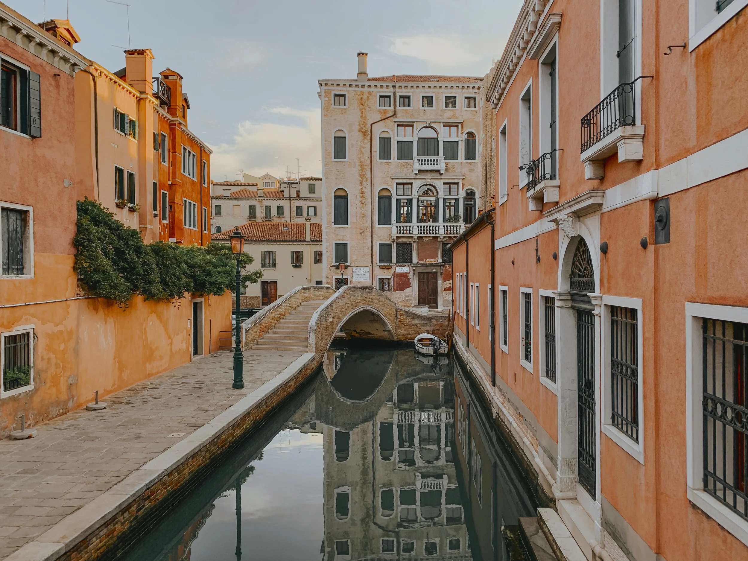 A narrow canal with calm water running between colorful old buildings in Venice, Italy. A small arched stone bridge crosses the canal, and a boat floats on the water. The buildings have windows with black iron railings and balconies, and the sky is partly cloudy.