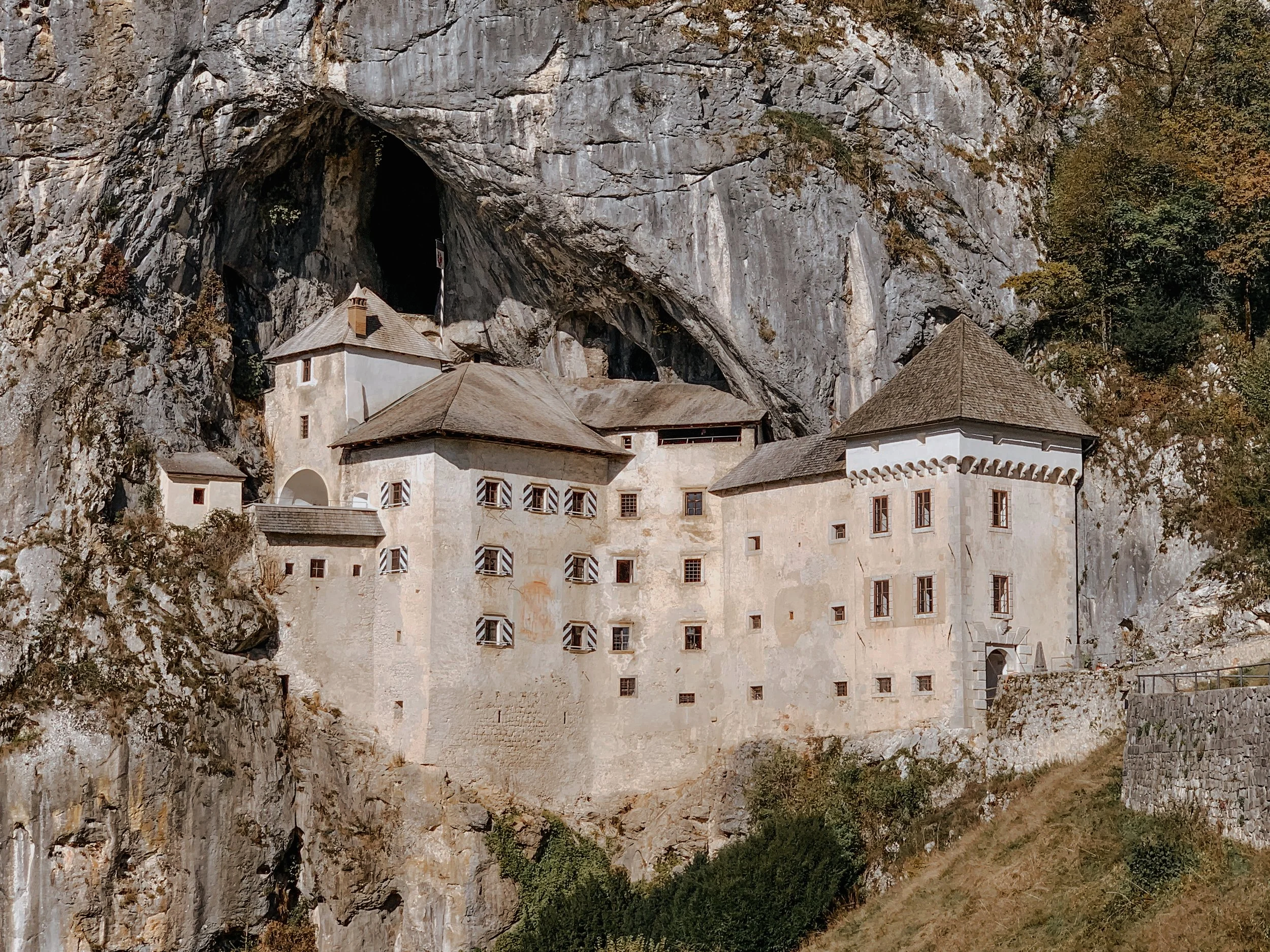 A medieval castle built into a mountainside with a large cave or opening above the building, surrounded by trees and rocky terrain.