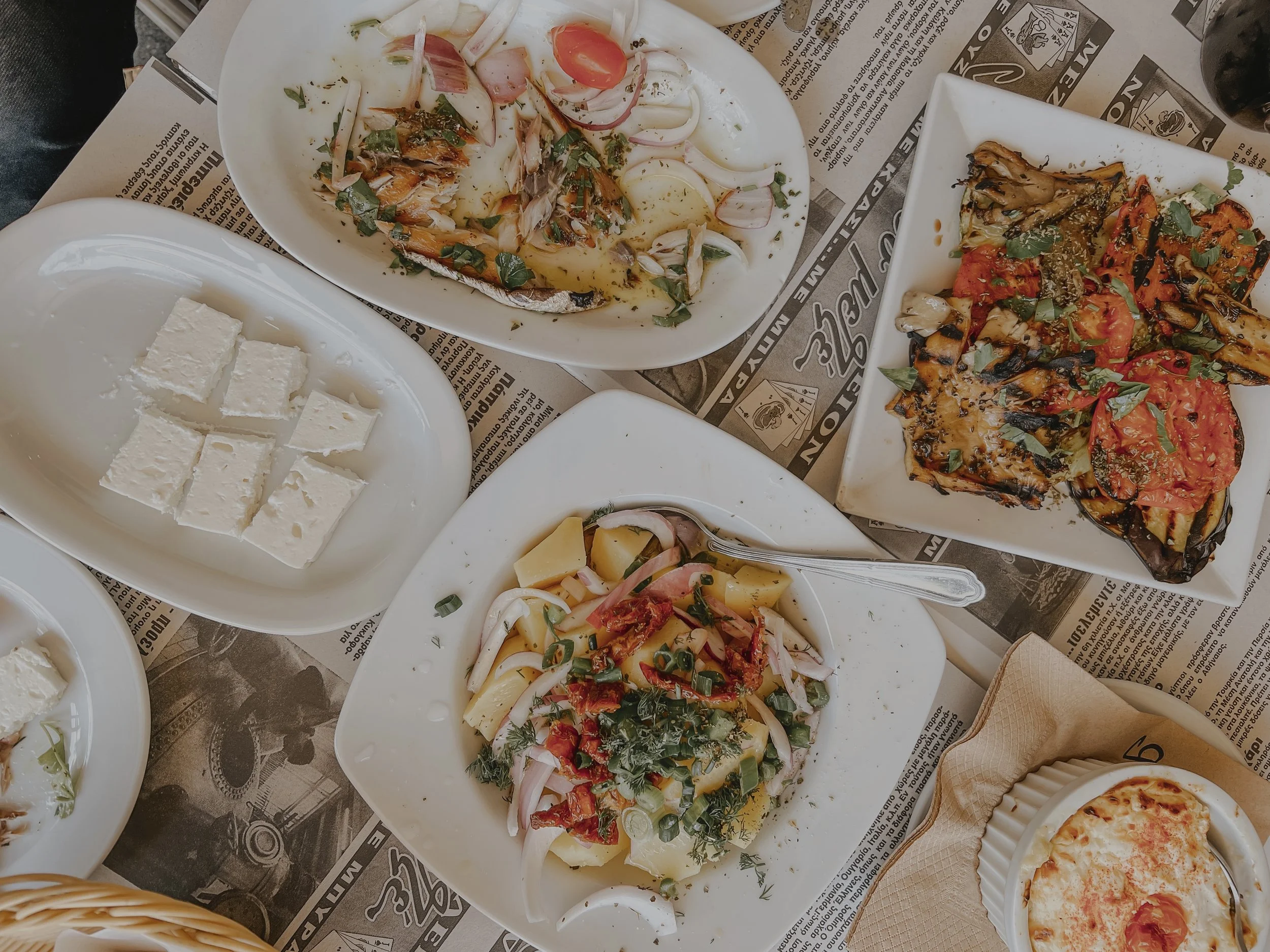 Assorted Mediterranean dishes on plates, including grilled vegetables with tomato, herbs, and balsamic glaze, a potato salad with herbs and onions, feta cheese, and a small baked dish topped with cheese on a paper-lined surface.