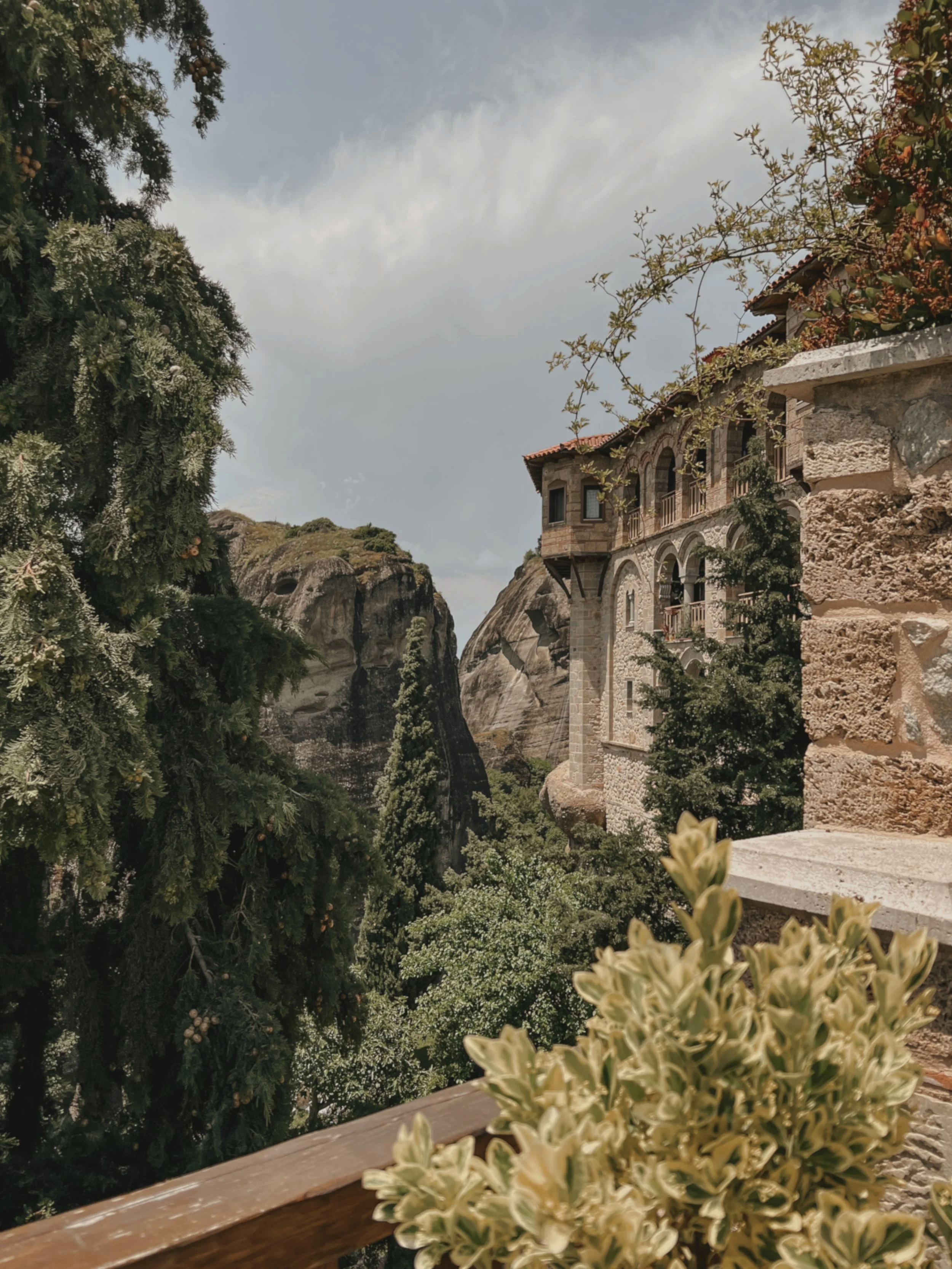 Mountain landscape with a stone building, greenery, trees, and rocky cliffs under a cloudy sky.