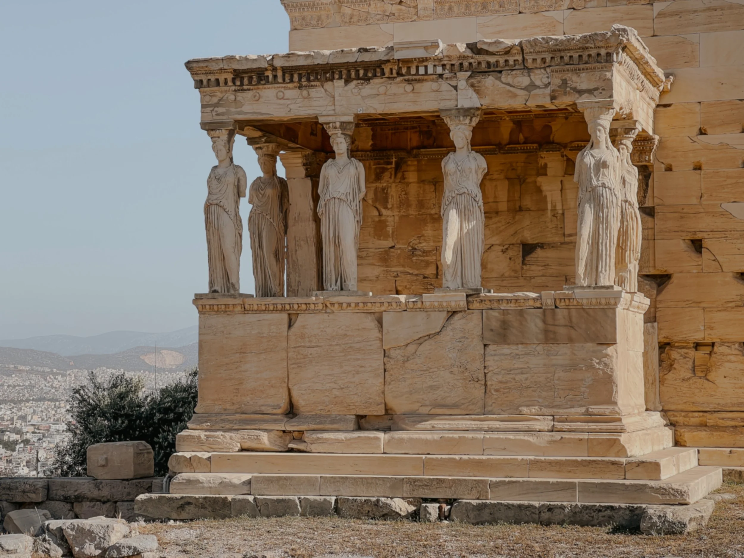Ancient Greek temple featuring six standing caryatids supporting the structure, with a cityscape and mountains in the background.