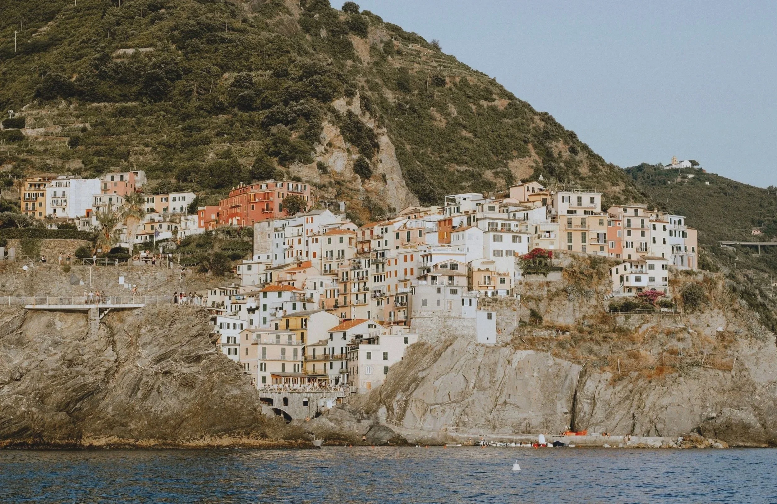 Colorful houses built on a steep hillside overlooking the water, with some boats visible near the rocky shoreline.
