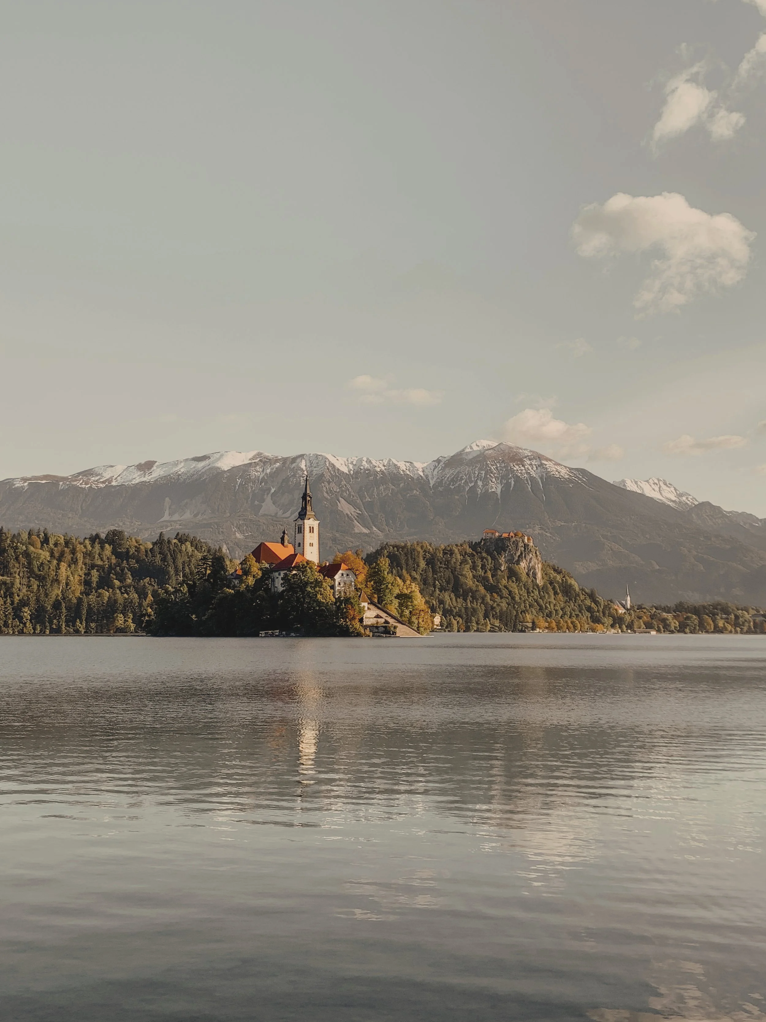 A church with a tall steeple on a small island in a lake, surrounded by lush trees and mountains with snow on the peaks in the background.