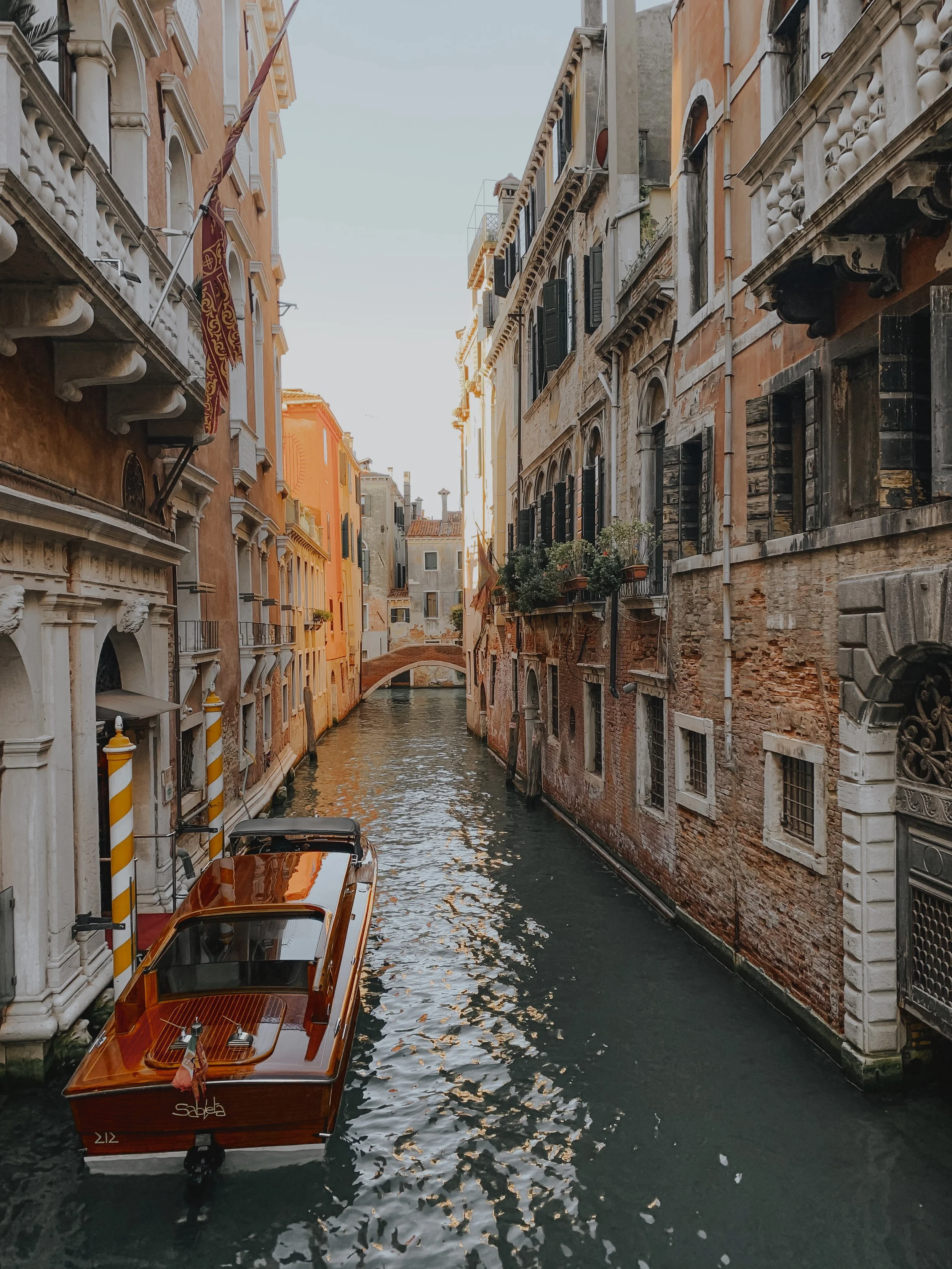A narrow canal in Venice, Italy, with a wooden boat floating next to old, colorful buildings with a small bridge in the background.