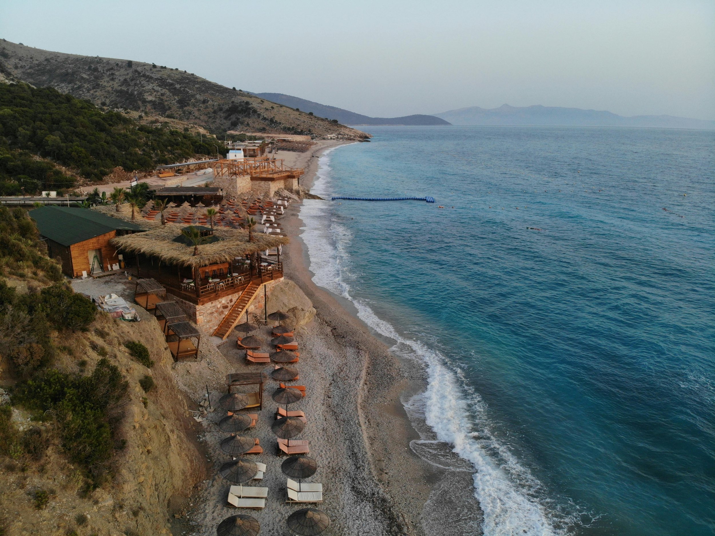 Aerial view of a beach resort with pebbled beach, thatched roof structures, sun loungers, and umbrellas, with clear blue water and mountains in the background.