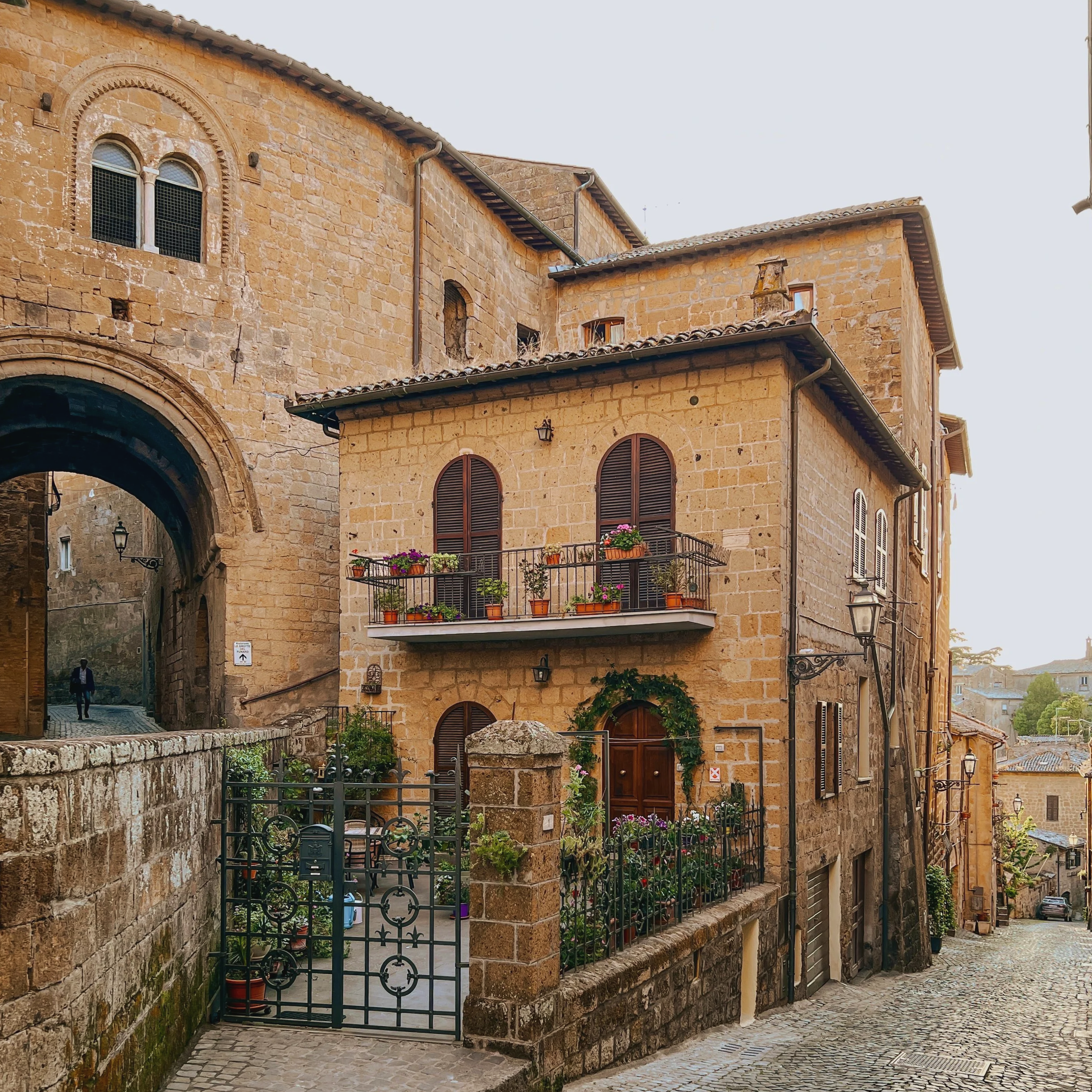 Stone building with arched windows and a balcony with flower pots, on a cobblestone street in a historic European town.