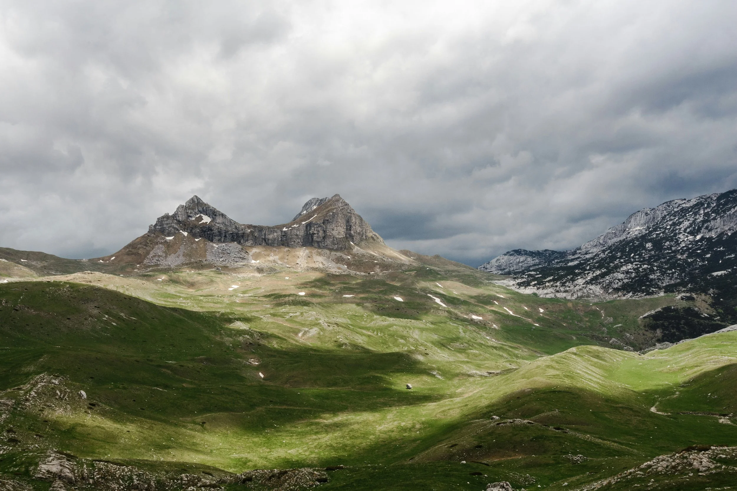 A mountainous landscape with green grassy hills and rocky peaks under a cloudy sky.