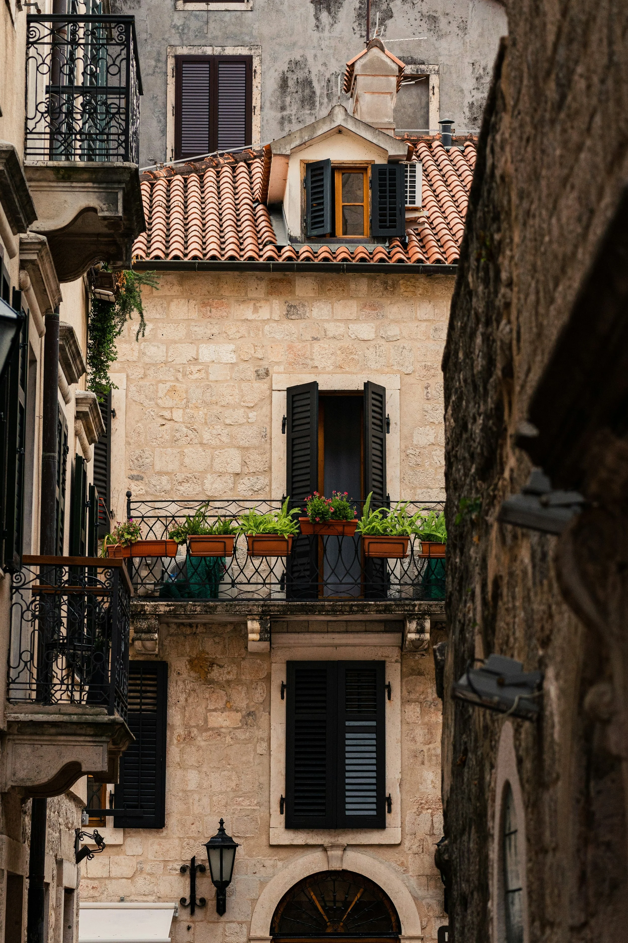 View of a stone building with black window shutters, a small balcony with potted plants, and a red-tiled roof in a European city.