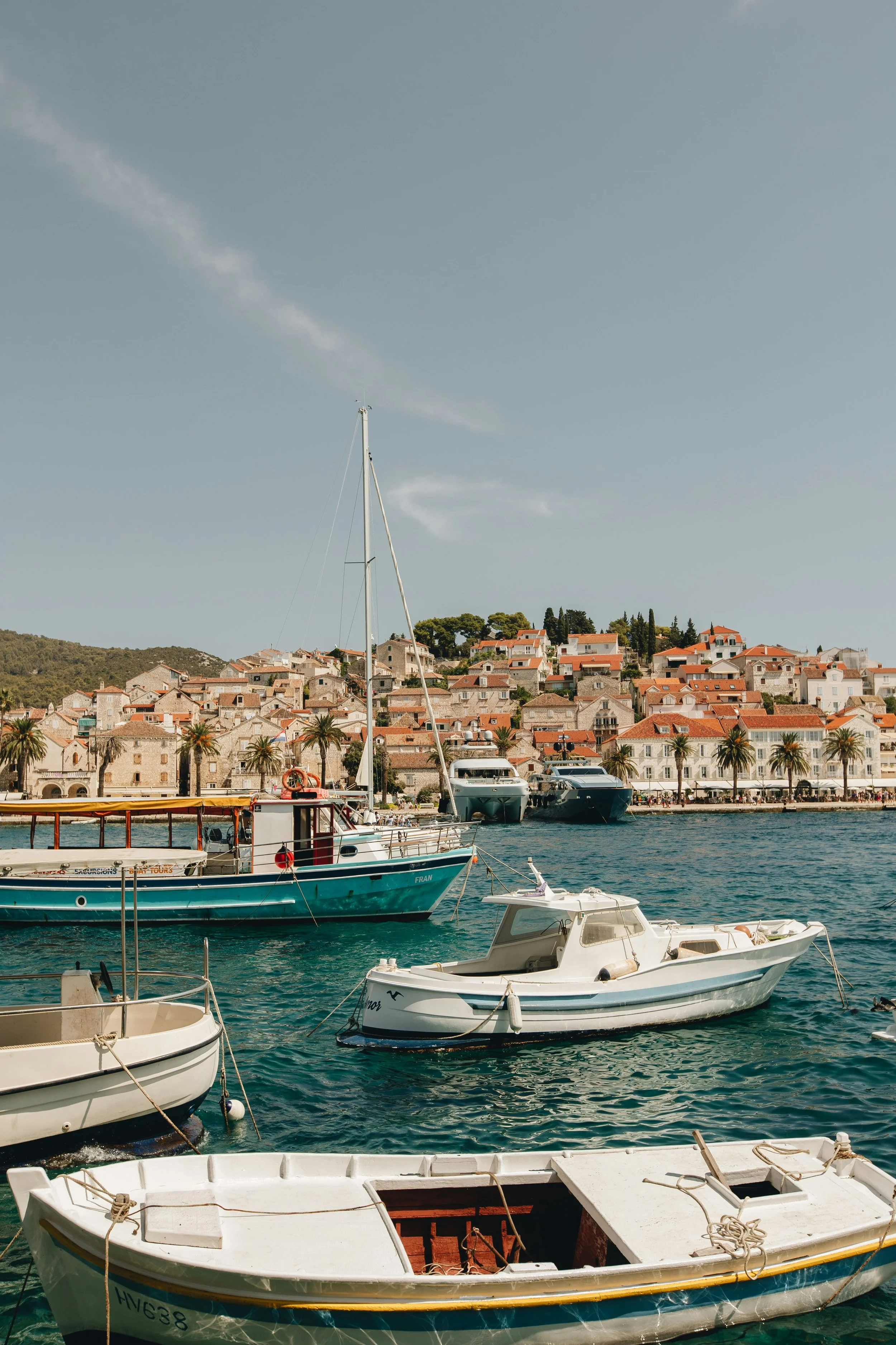 Boats docked in a harbor with a coastal town and hillside in the background.