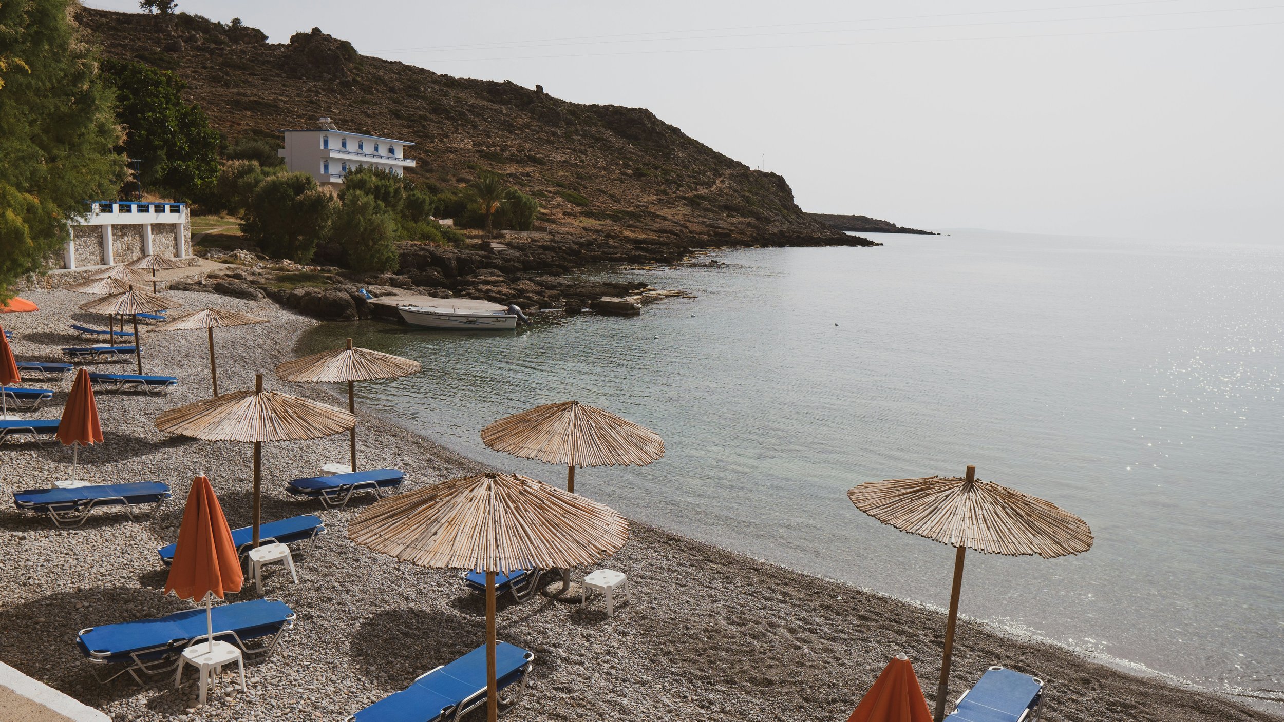 Empty beach with blue lounge chairs and straw umbrellas, calm water, hillside with white buildings, overcast sky.