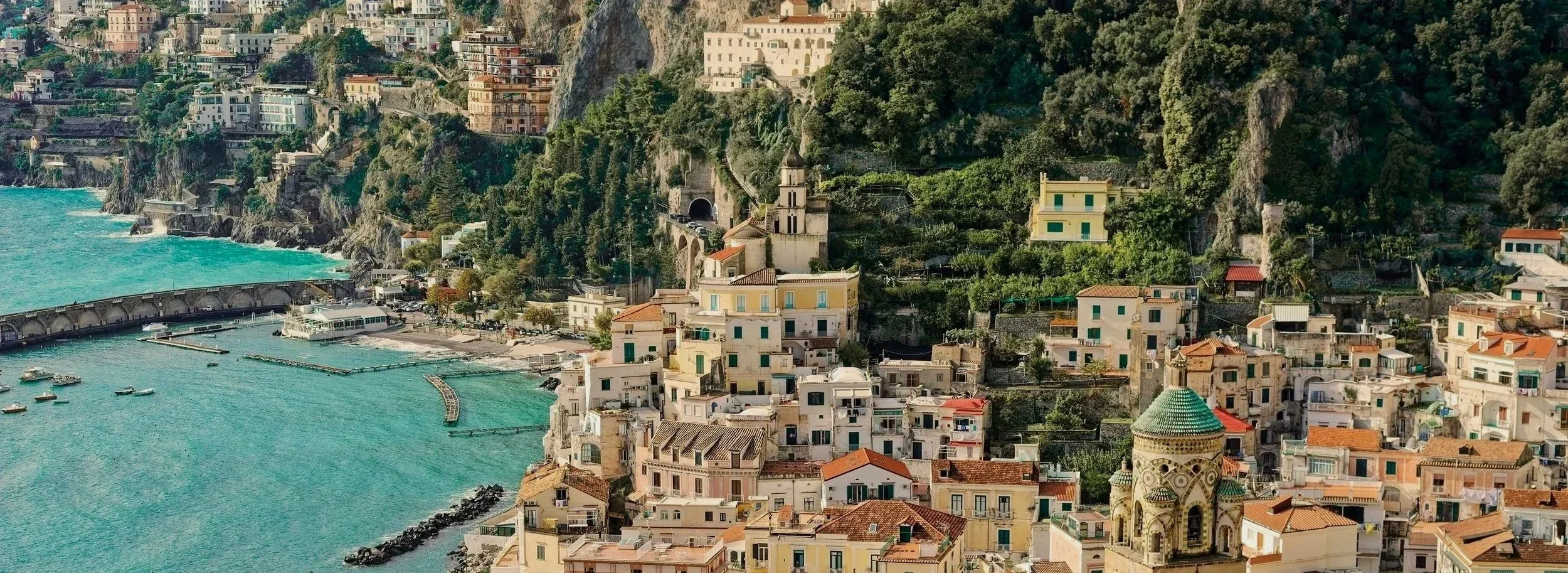 Coastal town built on steep cliffs with colorful buildings, a church with a green-tiled dome, and a turquoise harbor with boats.