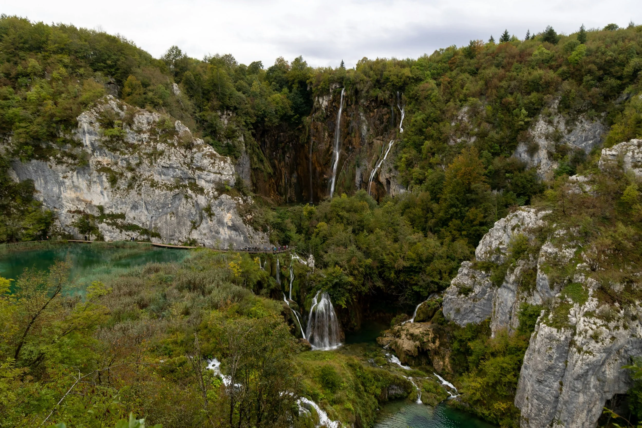Scenic view of a lush green canyon with waterfalls flowing into a turquoise lake, surrounded by dense trees and rocky cliffs.