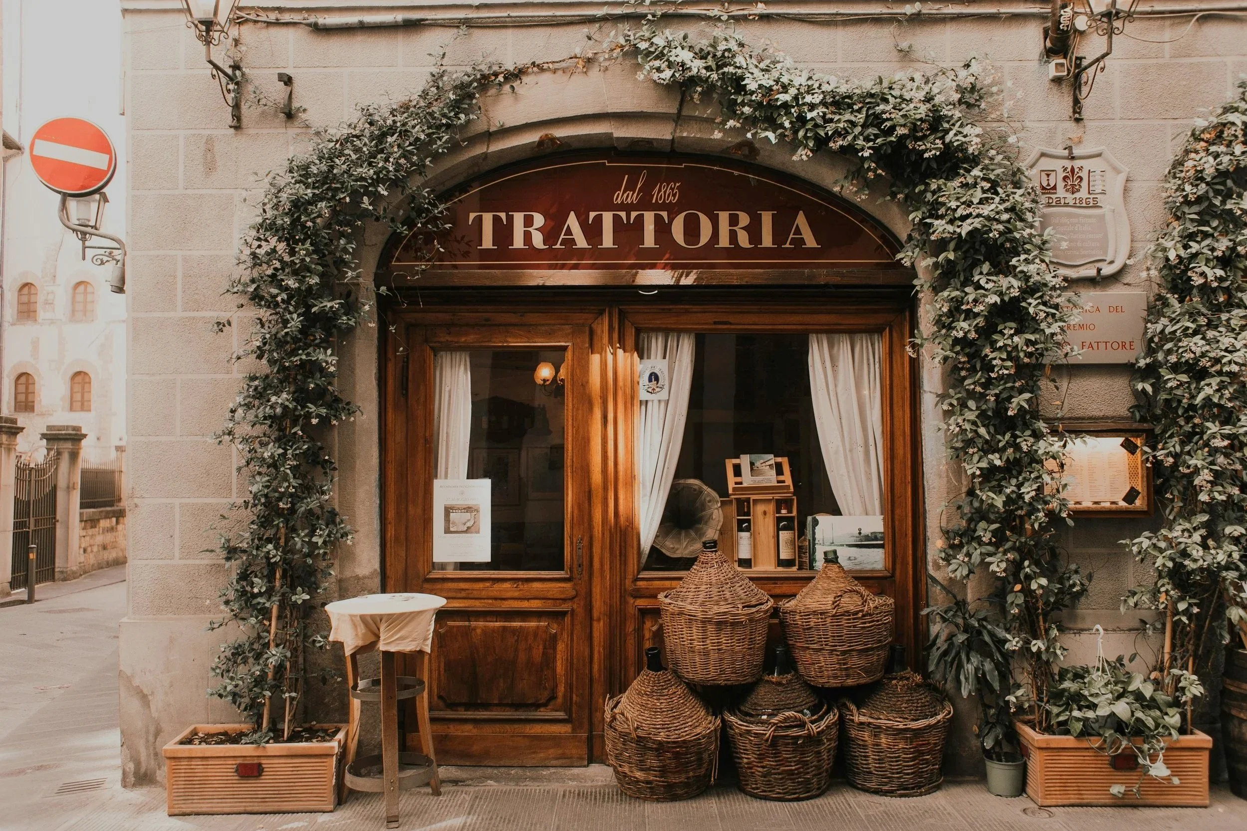 Exterior of a traditional Italian trattoria with wooden doors, potted plants, wicker baskets, and a sign reading 'dal 1885 Trattoria' above the entrance.