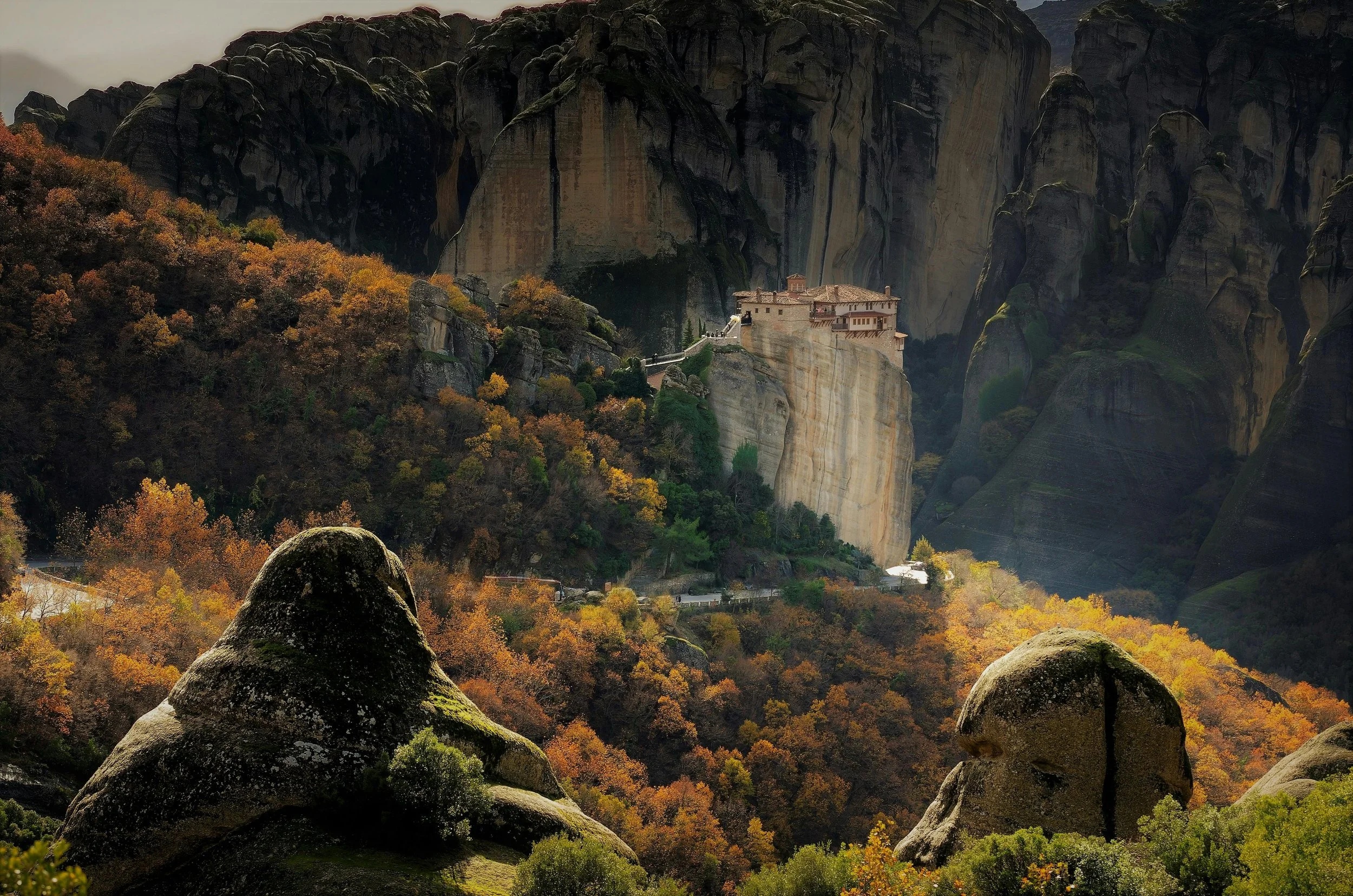 View of a monastery perched on a cliff in Meteora, Greece, surrounded by autumn-colored trees and large moss-covered rocks in the foreground.