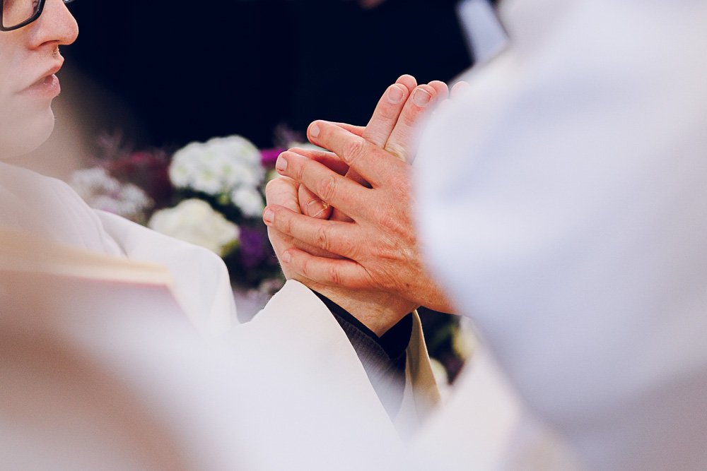 Eine Person, wahrscheinlich bei einer religiösen Zeremonie, hält die Hände einer anderen Person in Gebetshaltung. Im Hintergrund sind Blumen sichtbar.