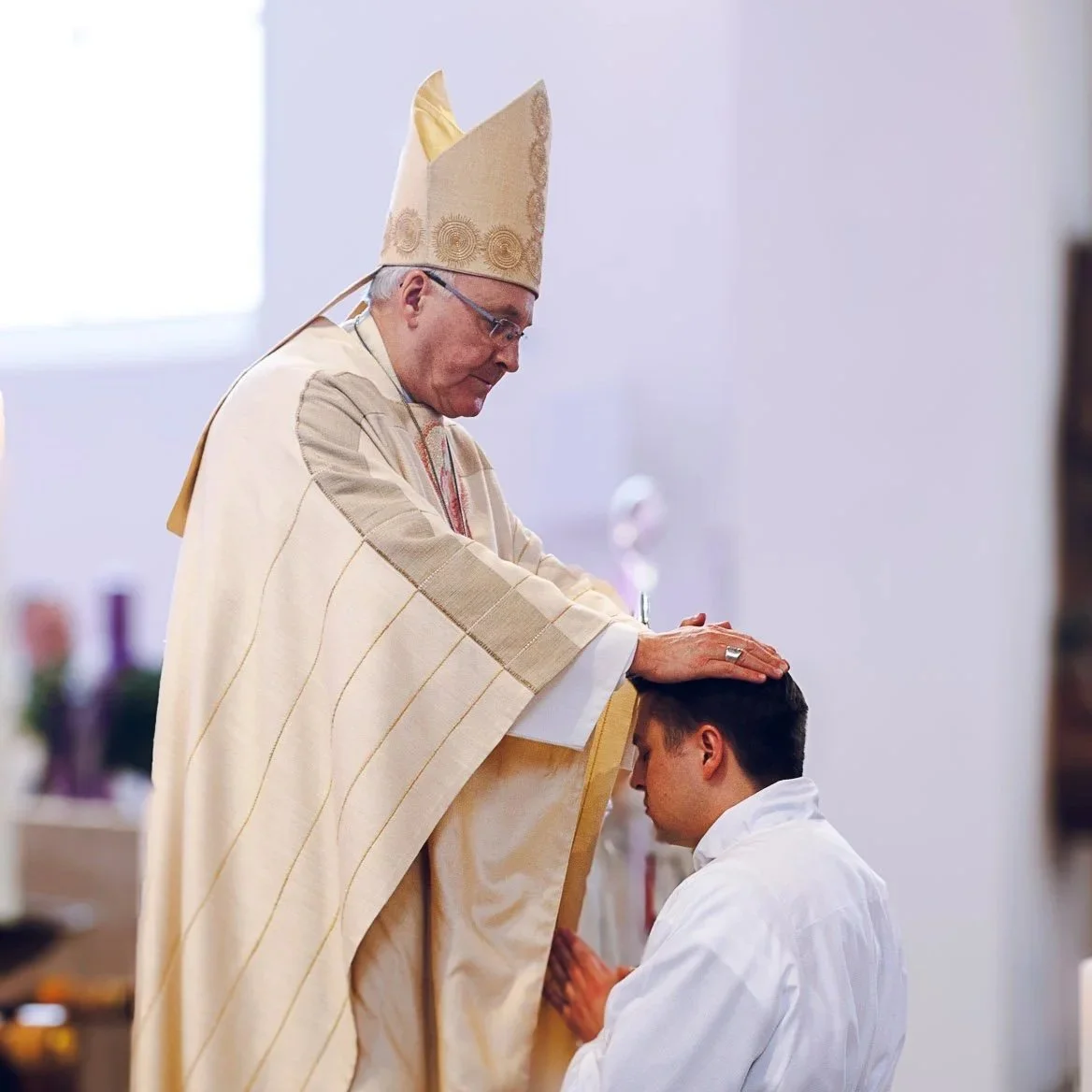 Ein Priester in goldener Kleidung segnet einen jungen Mann, der in weißem Gewand knieend mit gefalteten Händen betet, in einer Kirche mit Blumen und anderen Personen im Hintergrund.
