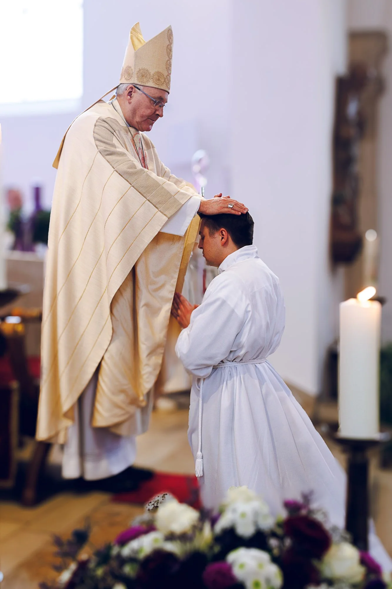Ein Priester in goldener Kleidung segnet einen jungen Mann, der in weißem Gewand knieend mit gefalteten Händen betet, in einer Kirche mit Blumen und anderen Personen im Hintergrund.