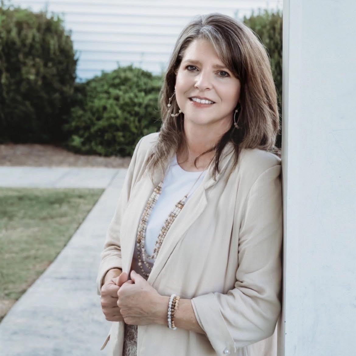 A woman with shoulder-length brown hair, wearing a beige blazer and jewelry, leaning against a white wall outside, smiling at the camera.
