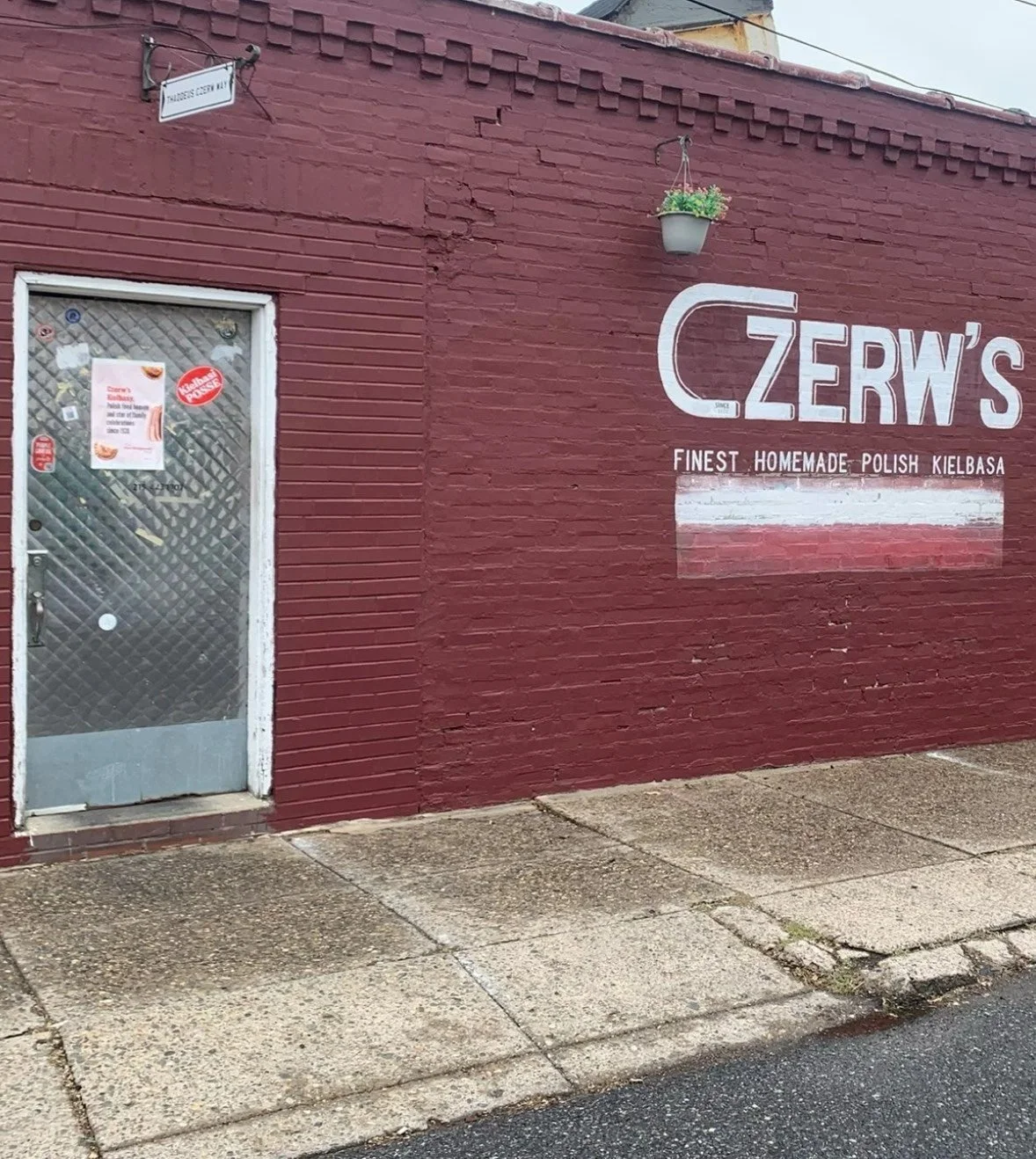 Red brick wall with a door on the left and a painted sign on the right reading 'CZERW'S FINEST HOMEMADE POLISH KIELBASA'. There's a small potted plant hanging above the sign and a street sign in the top left corner that says 'WASHBURN CERRY ST'.
