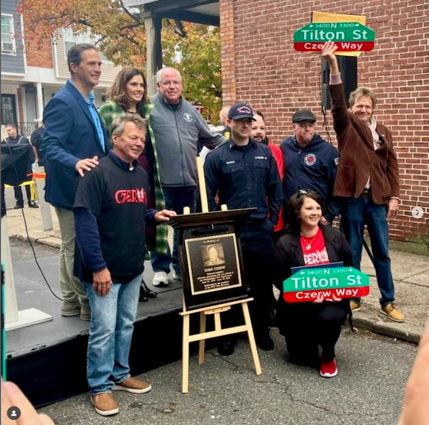 Group of people gathered outdoors for a street dedication ceremony, with one woman holding a street sign for Tilton Street and others standing around a plaque on an easel.
