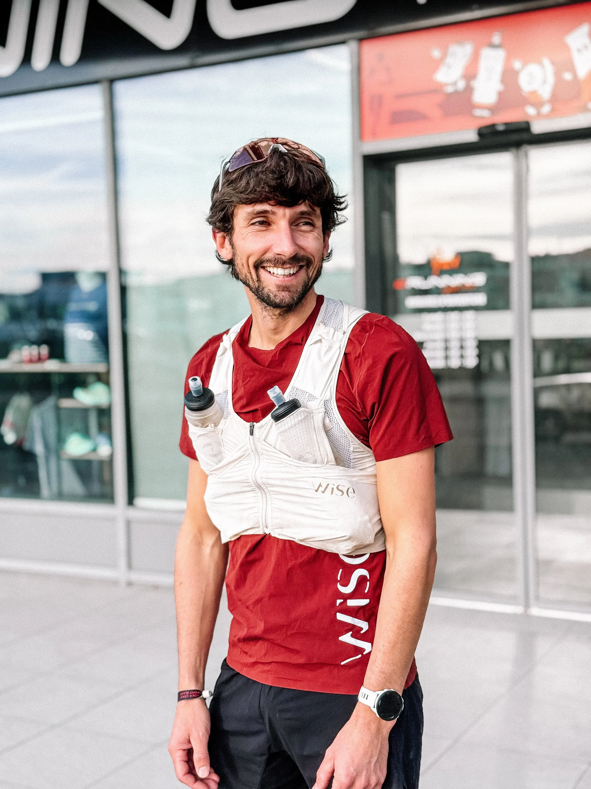 Un homme souriant portant un t-shirt rouge, une montre blanche, et un gilet dorsal avec des bouteilles d'eau, se trouvant devant une entrée de magasin.