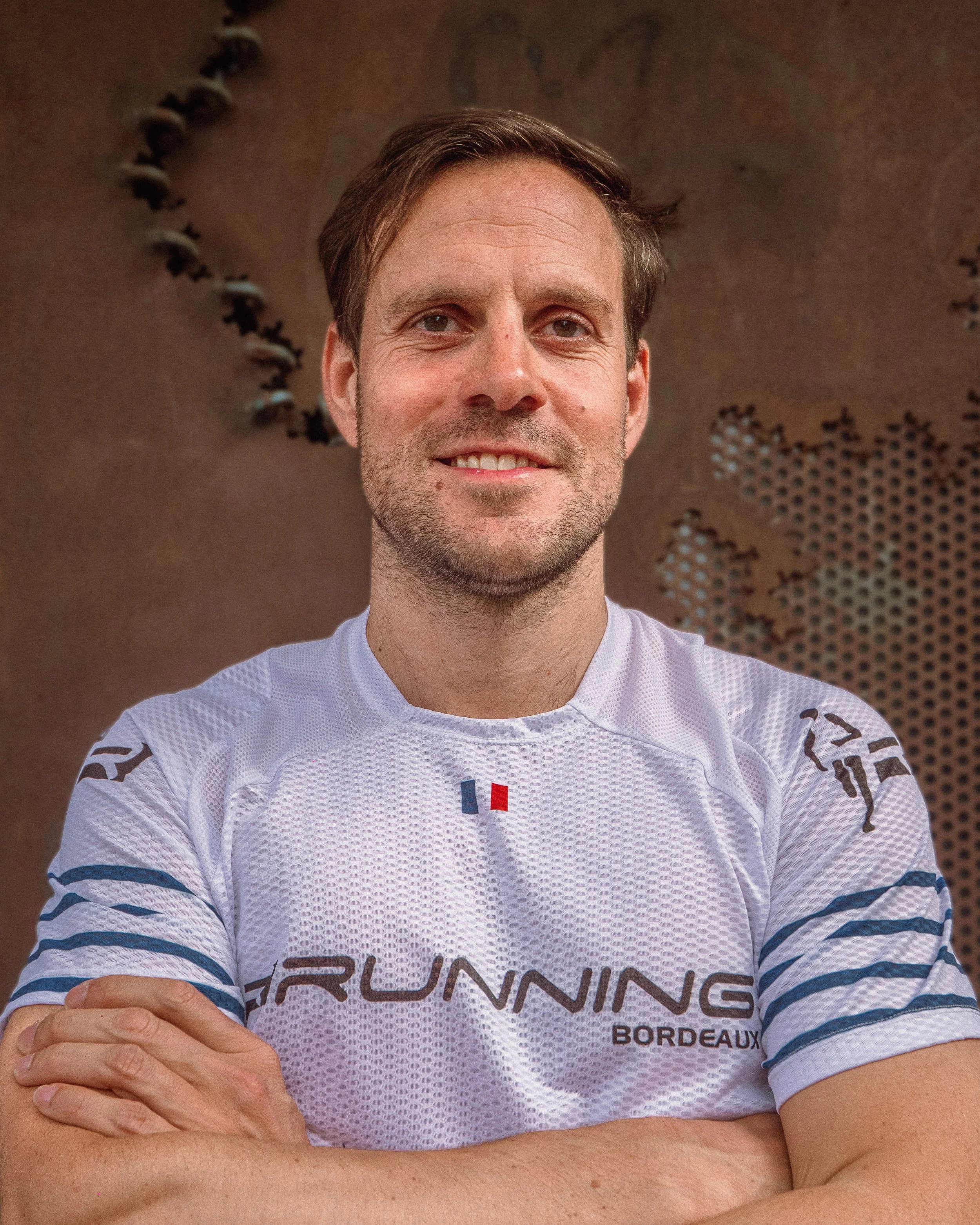 Homme souriant en maillot de sport blanc avec écrit 'Running Bordeaux', bras croisés, debout devant une sculpture ou un mur métallique.